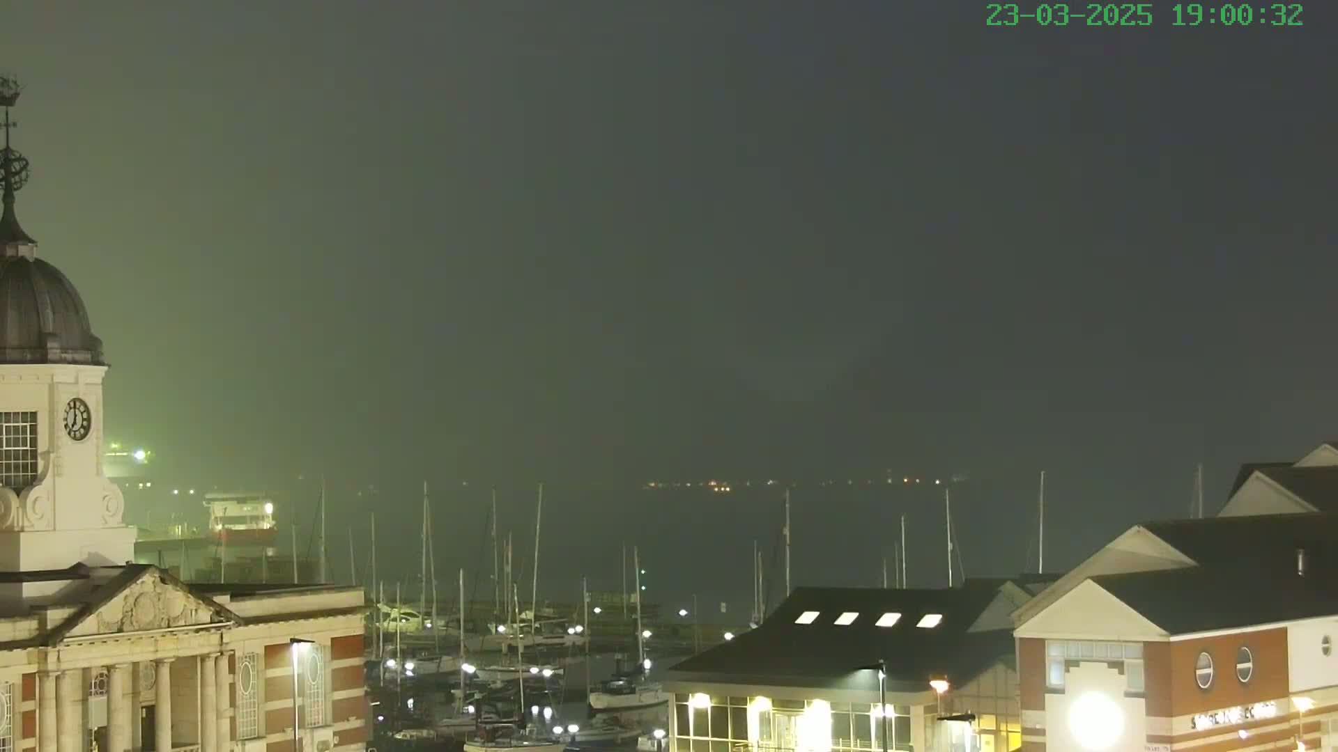 A nighttime view of a harbor filled with boats under a foggy sky, with a clock tower building in the foreground.