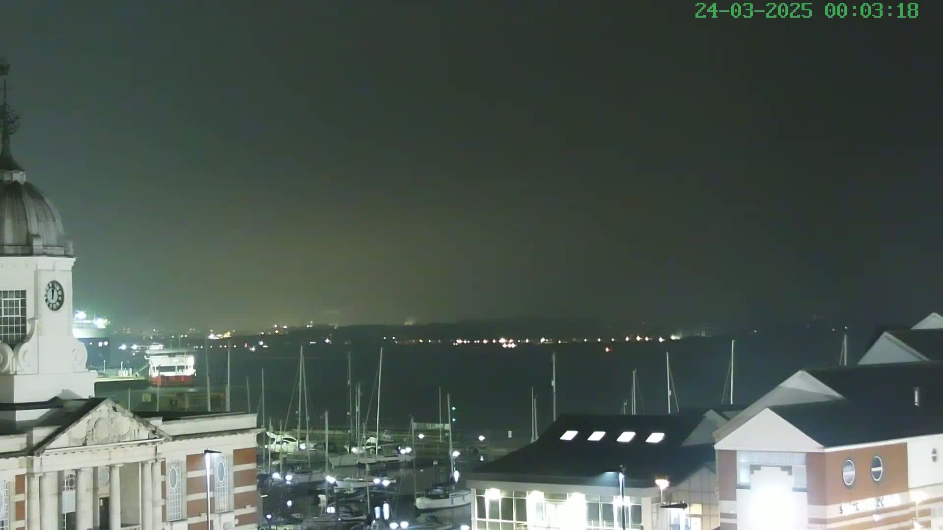 A nighttime view of a harbor with numerous boats docked, a clock tower, and buildings under a dark, hazy sky.