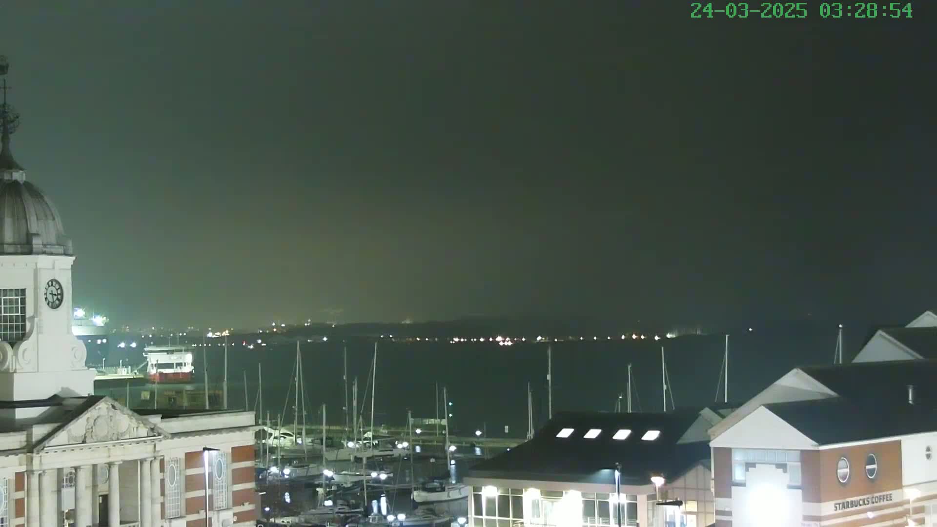 A nighttime view of a harbor filled with sailboats under a dark, hazy sky, with a clock tower and buildings visible in the foreground.