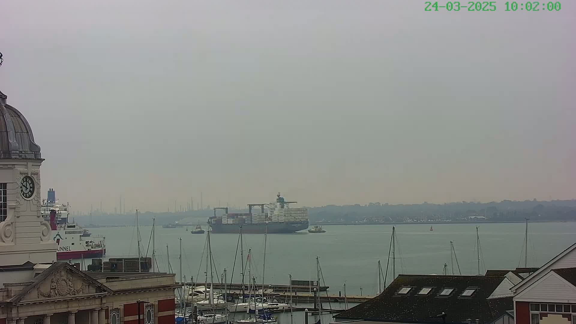 A large container ship is visible on a hazy, gray day, sailing past a harbor filled with moored sailboats and a building with a clock tower.