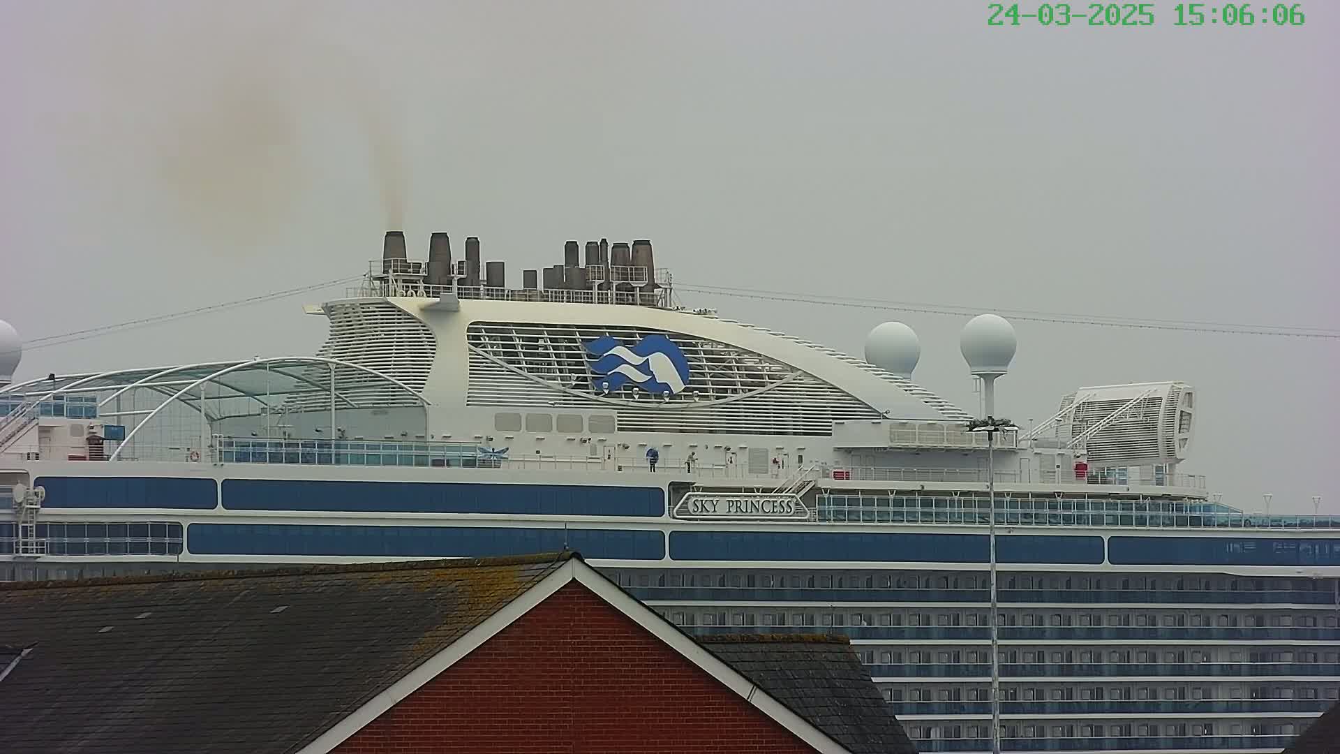 The upper deck of a large cruise ship is visible, emitting a plume of smoke under an overcast sky.