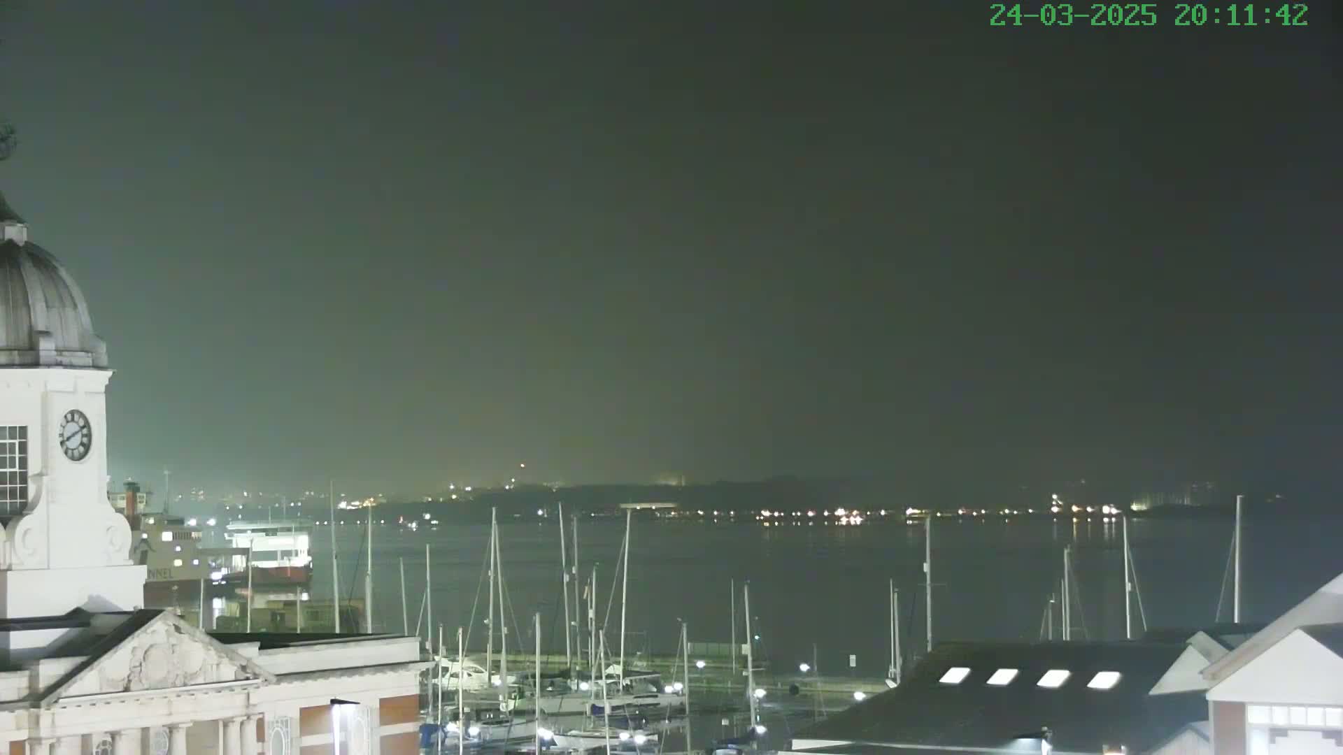 A nighttime view of a harbor filled with docked sailboats, seen from a building with a clock tower, under a hazy sky.