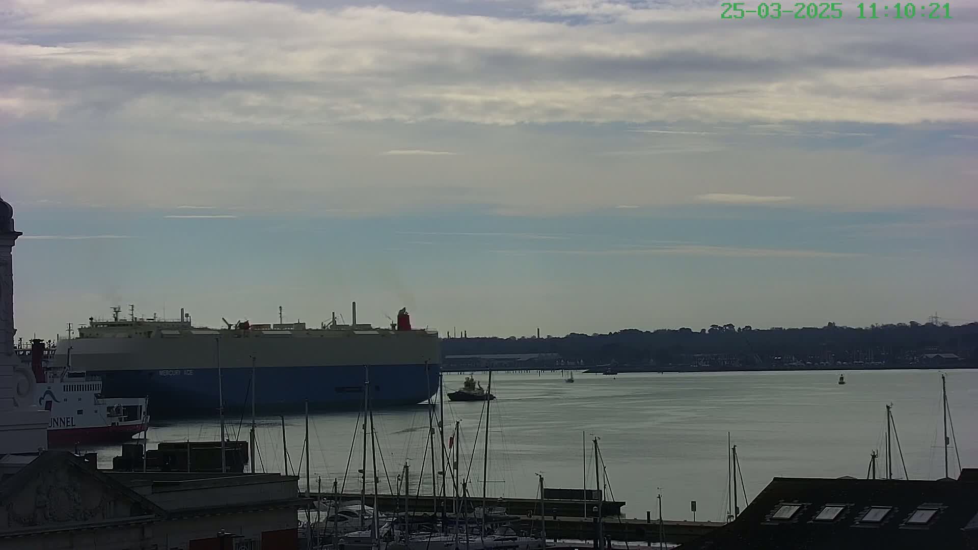 A large car carrier ship and a smaller tugboat are visible in a harbor under a mostly cloudy sky.
