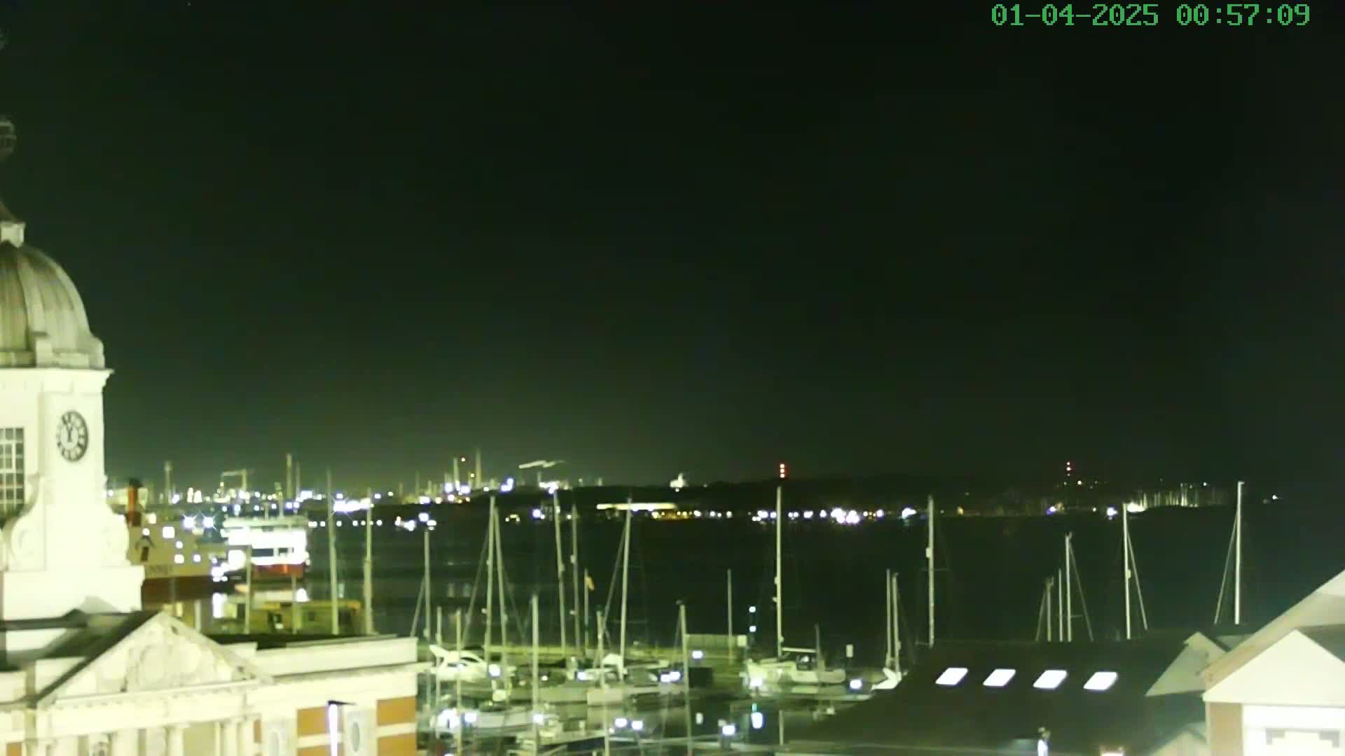 A nighttime view from atop a building shows a marina filled with docked sailboats, with a city skyline and industrial area illuminated in the distance under a clear, dark sky.