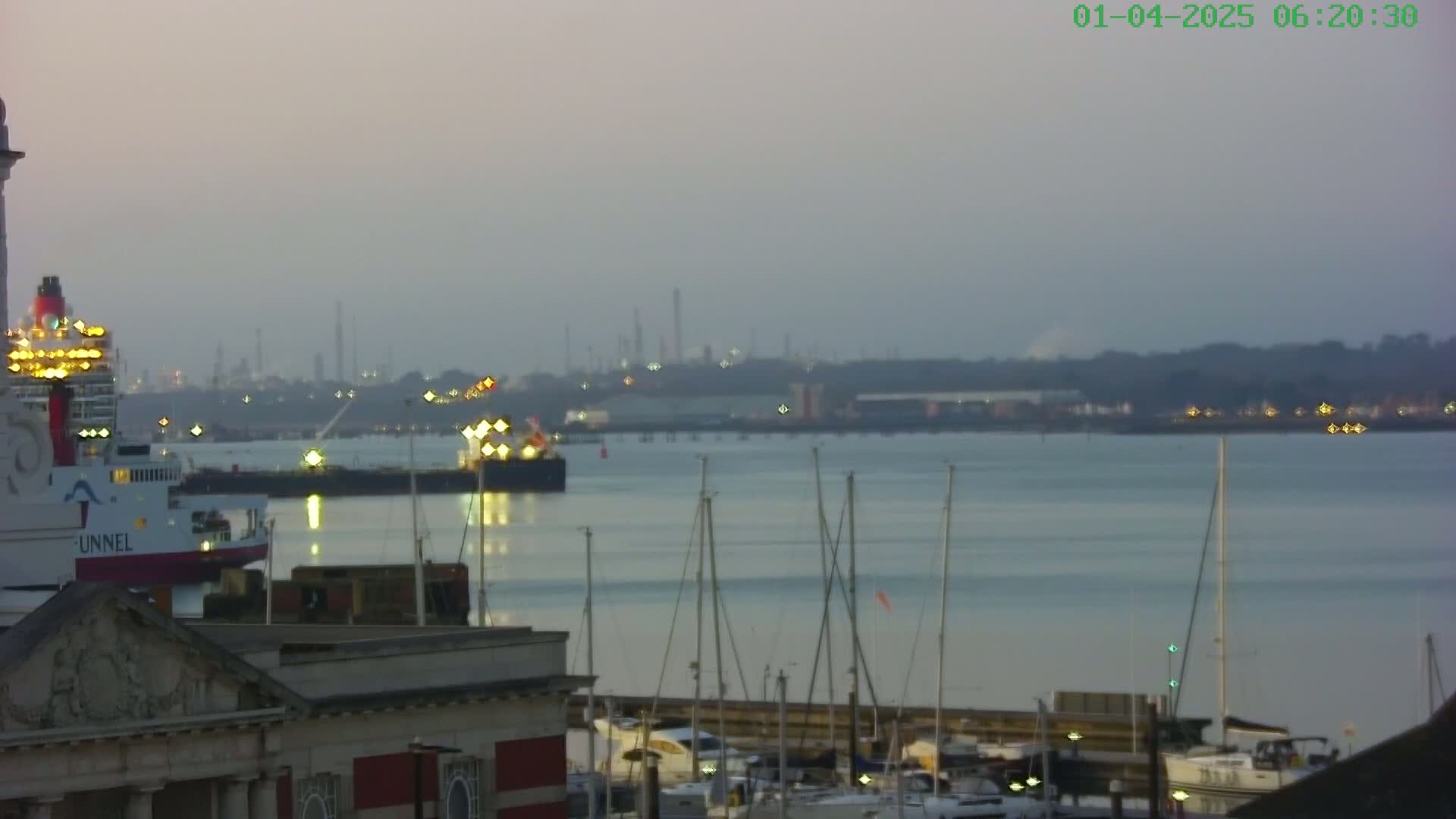 A large ferry is docked near a harbor filled with sailboats at dawn under a hazy sky, with an industrial area visible in the distance.