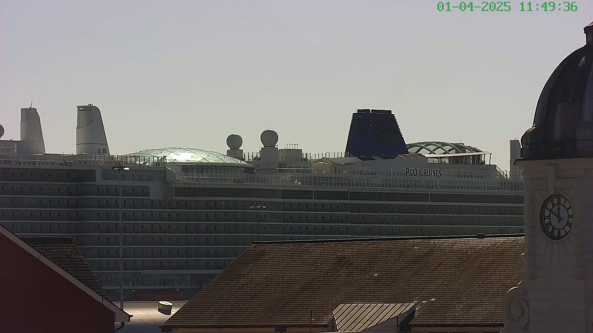 A large cruise ship is visible in the distance under a hazy sky, partially obscured by the roofs of nearby buildings, one of which is a clock tower.
