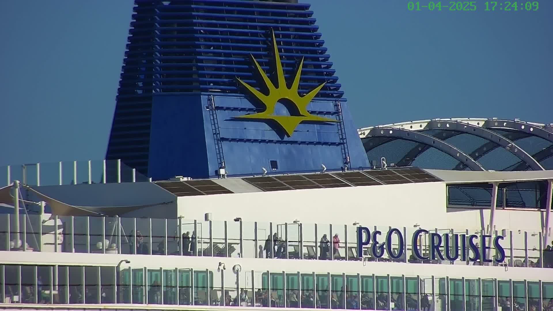 A P&O Cruises ship's deck, under a clear blue sky, shows people on an upper deck with glass railings and a blue smokestack featuring the company's logo.