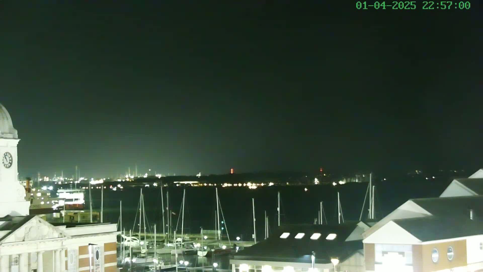 A nighttime view of a harbor filled with sailboats, a cityscape in the distance, and a portion of a clock tower in the foreground under a dark, clear sky.