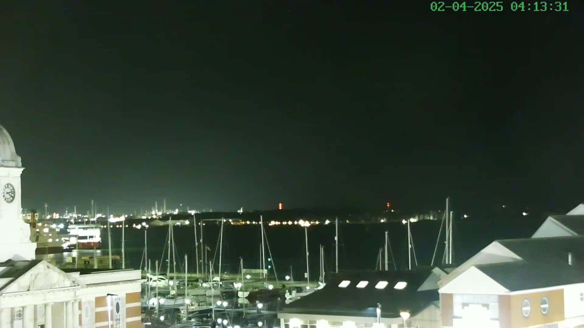 A nighttime view of a harbor filled with docked sailboats, with a clock tower and buildings in the foreground under a clear, dark sky.