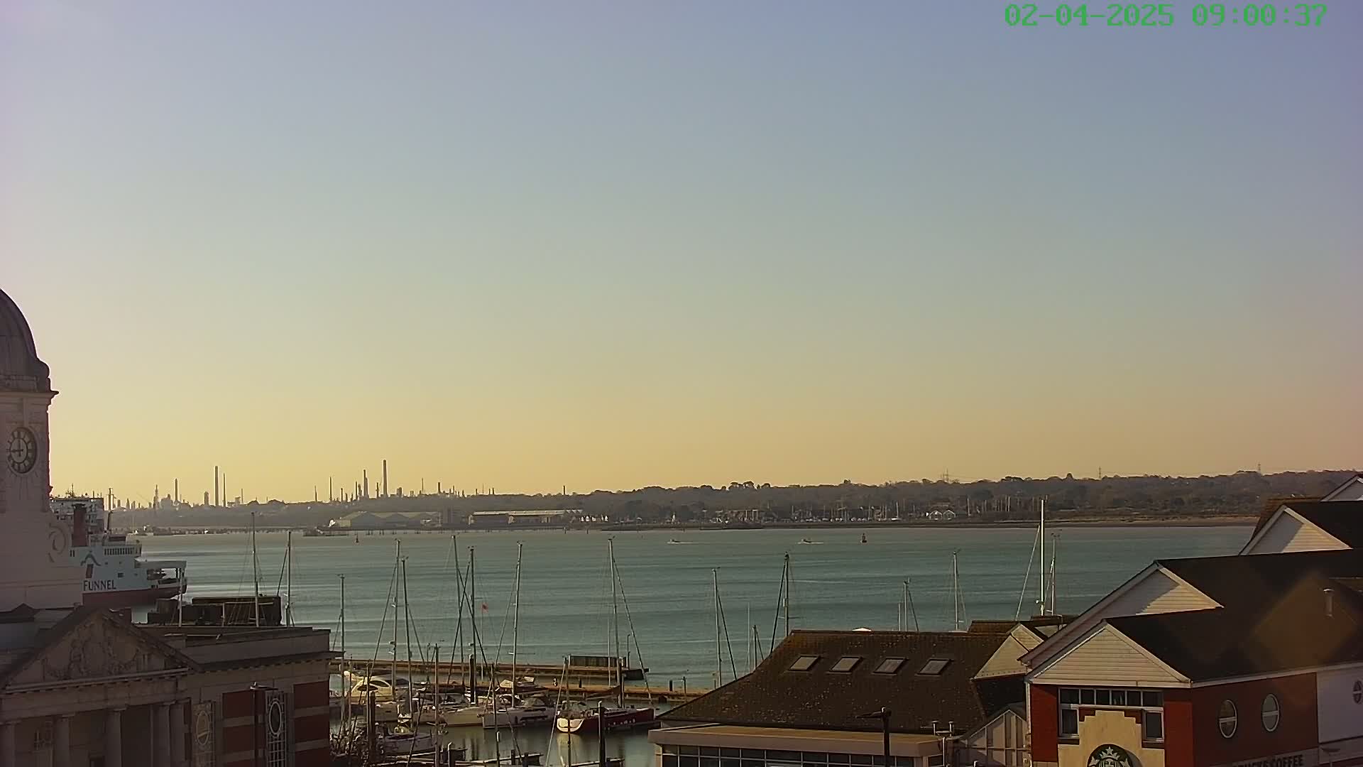 A calm body of water with numerous sailboats docked near buildings, with an industrial skyline visible in the distance under a clear, pale sky.