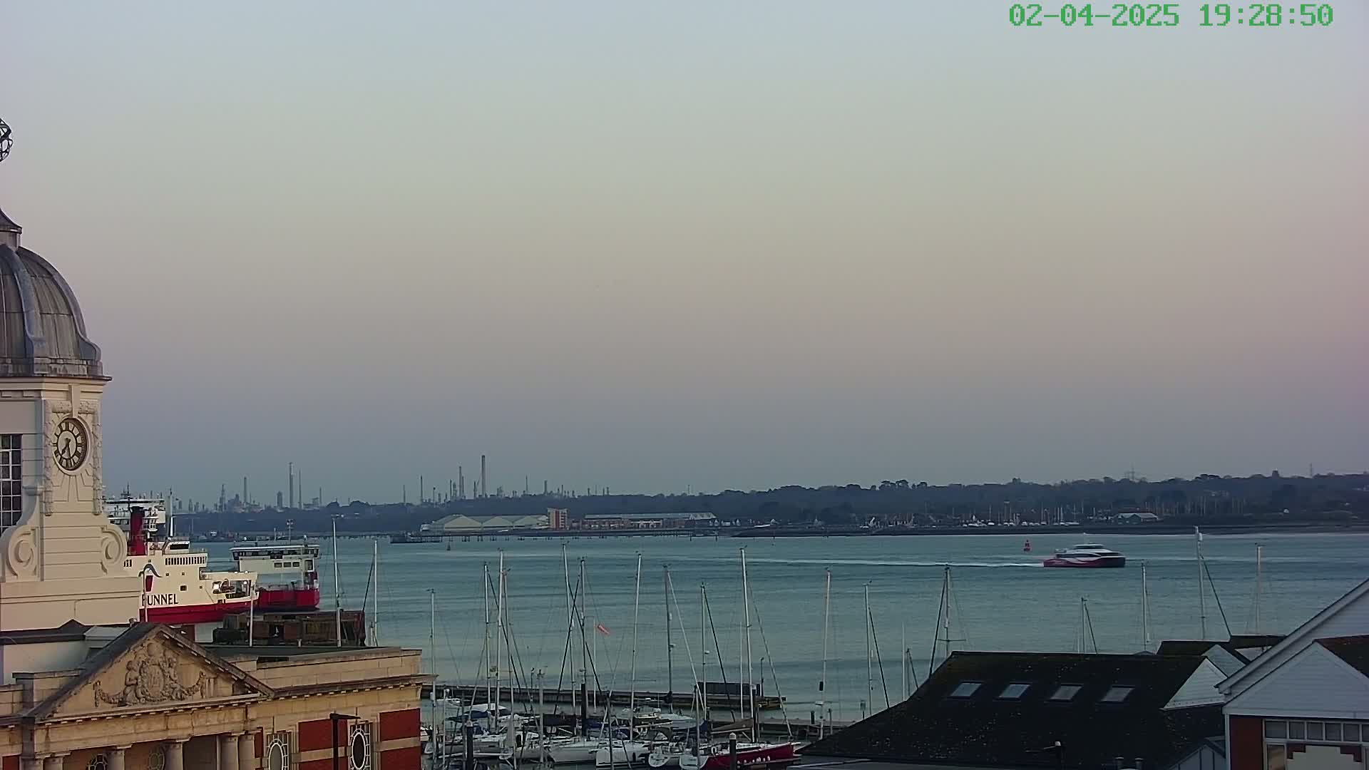 A calm, dusky waterfront scene shows a clock tower, a ferry, numerous docked sailboats, and an industrial facility in the distance.