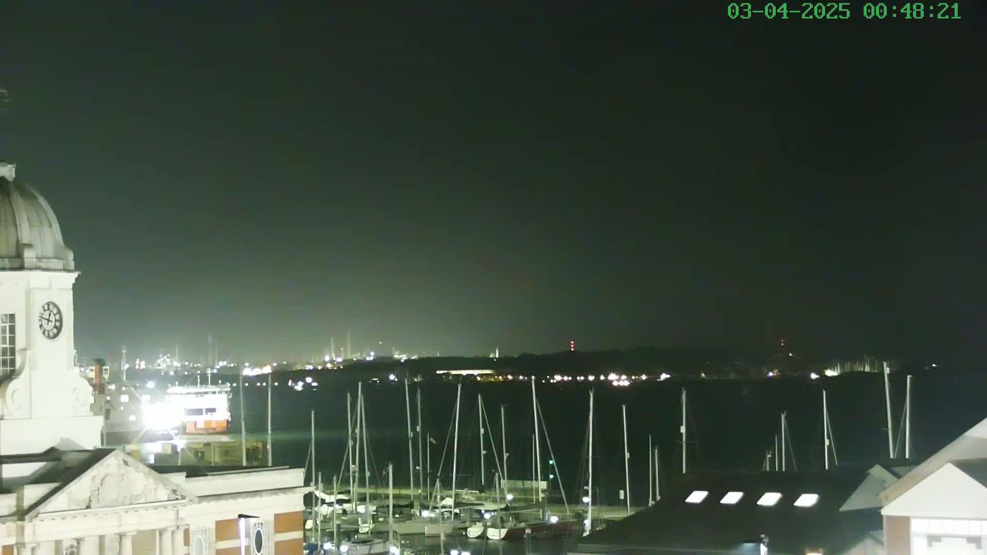 A nighttime view from a building with a clock tower overlooking a marina filled with sailboats and a distant, illuminated shoreline under a dark sky.