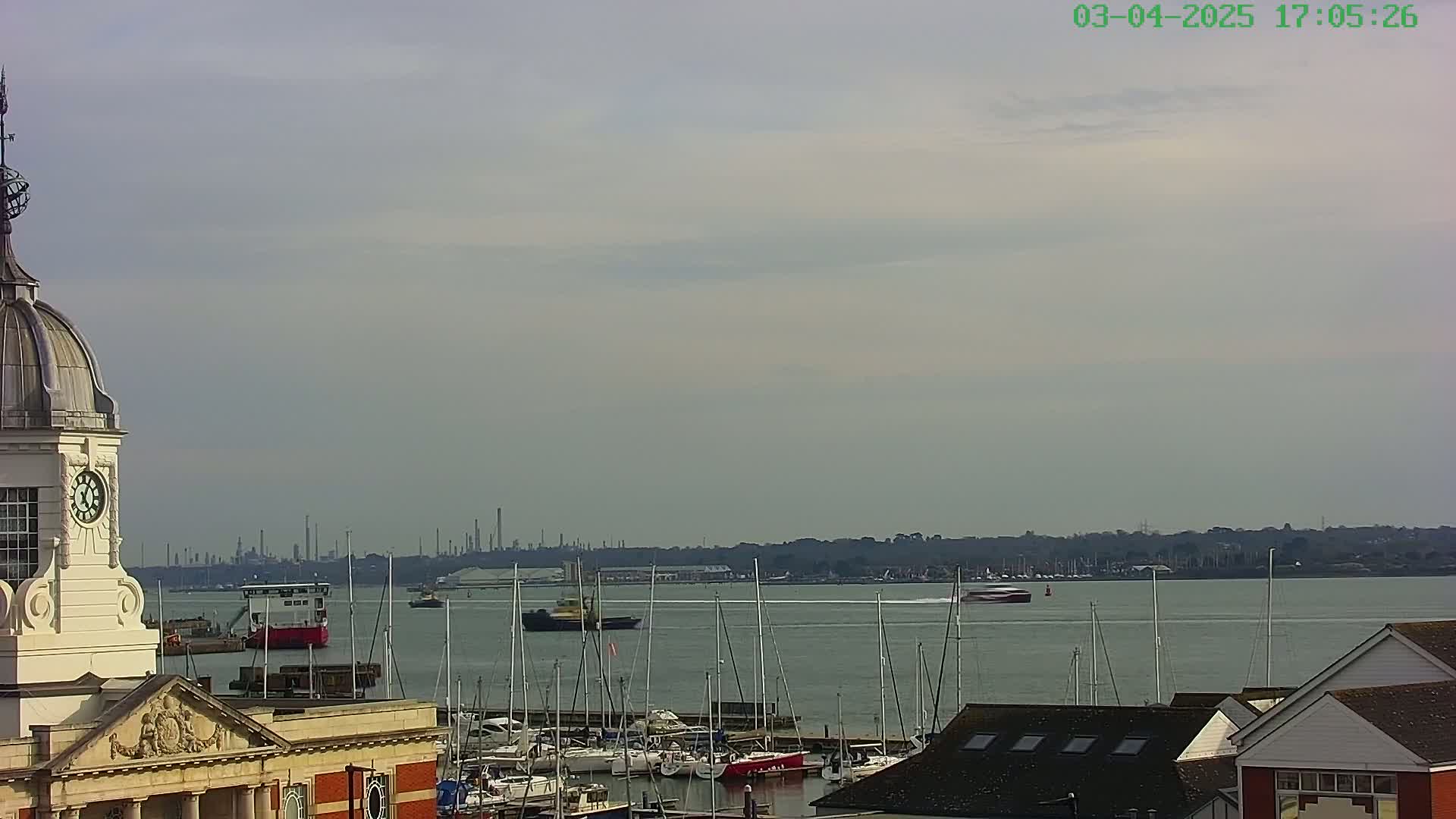 A clock tower overlooks a harbor filled with sailboats and motorboats, with a distant industrial skyline visible under a mostly cloudy sky.