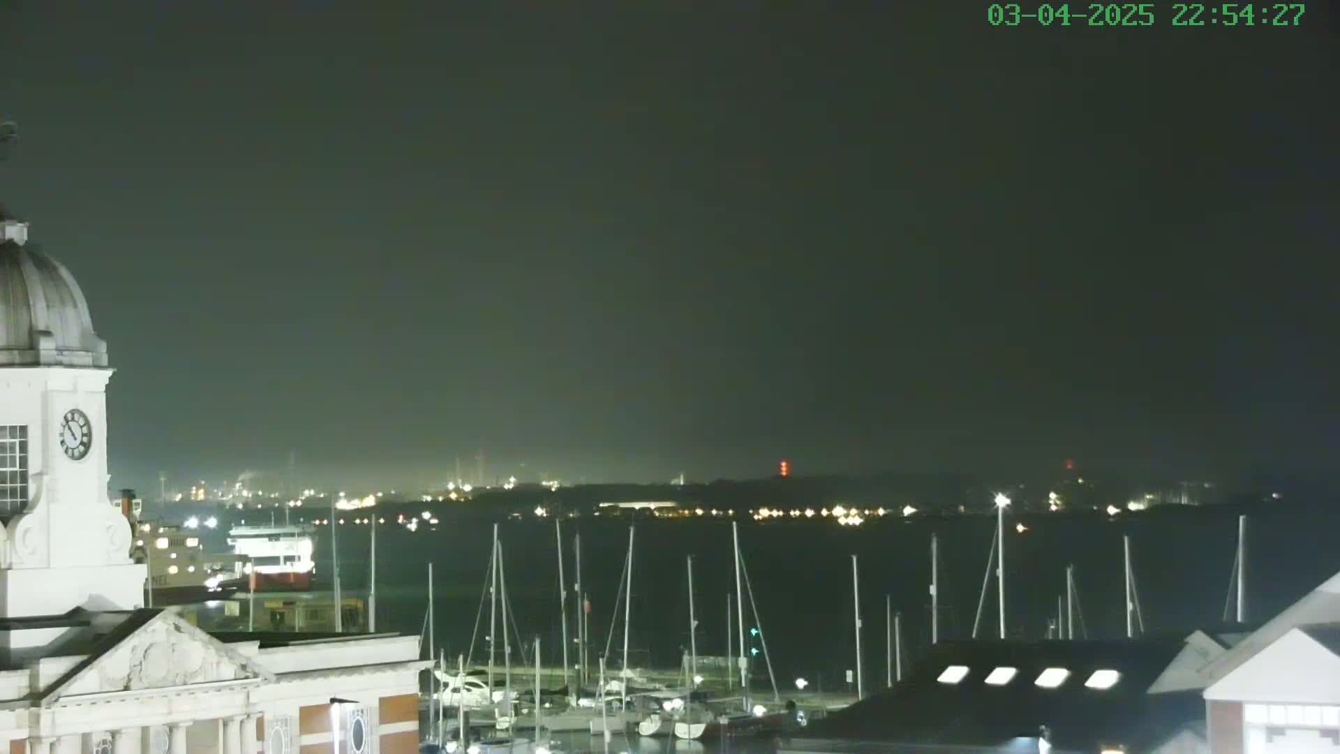 A clock tower overlooks a marina filled with sailboats at night under a dark, overcast sky.