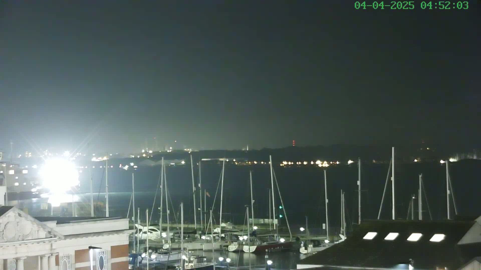 A nighttime view of a marina filled with sailboats, under a hazy, dark sky, with a distant shoreline faintly illuminated.