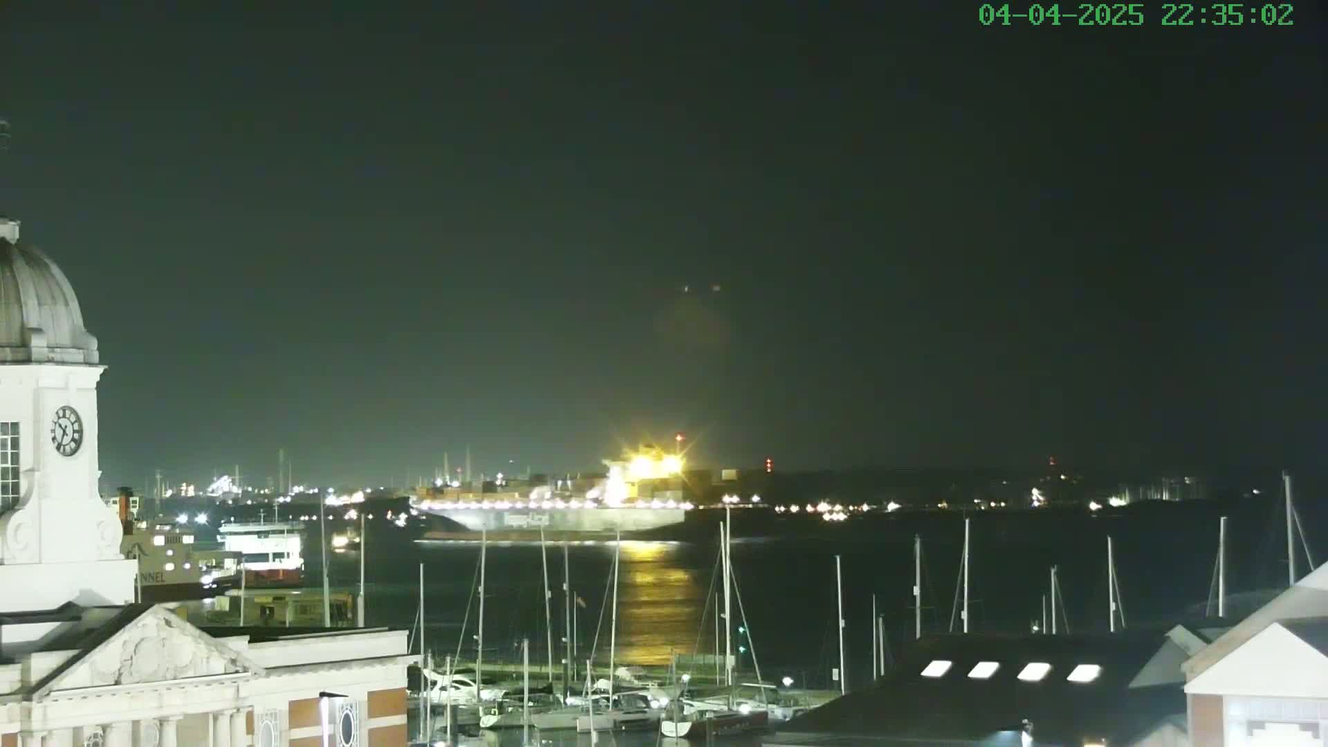 A large container ship is seen on a dark, calm body of water at night, illuminated by its own lights, with a marina of docked sailboats in the foreground and a clock tower to the left.