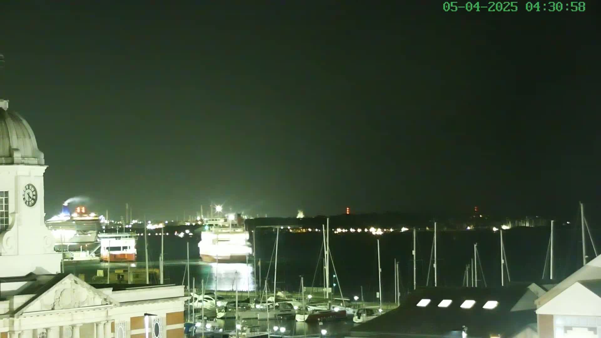 At night, a harbor scene shows a large ship and many sailboats docked, with a white building with a dome in the foreground under a dark sky.