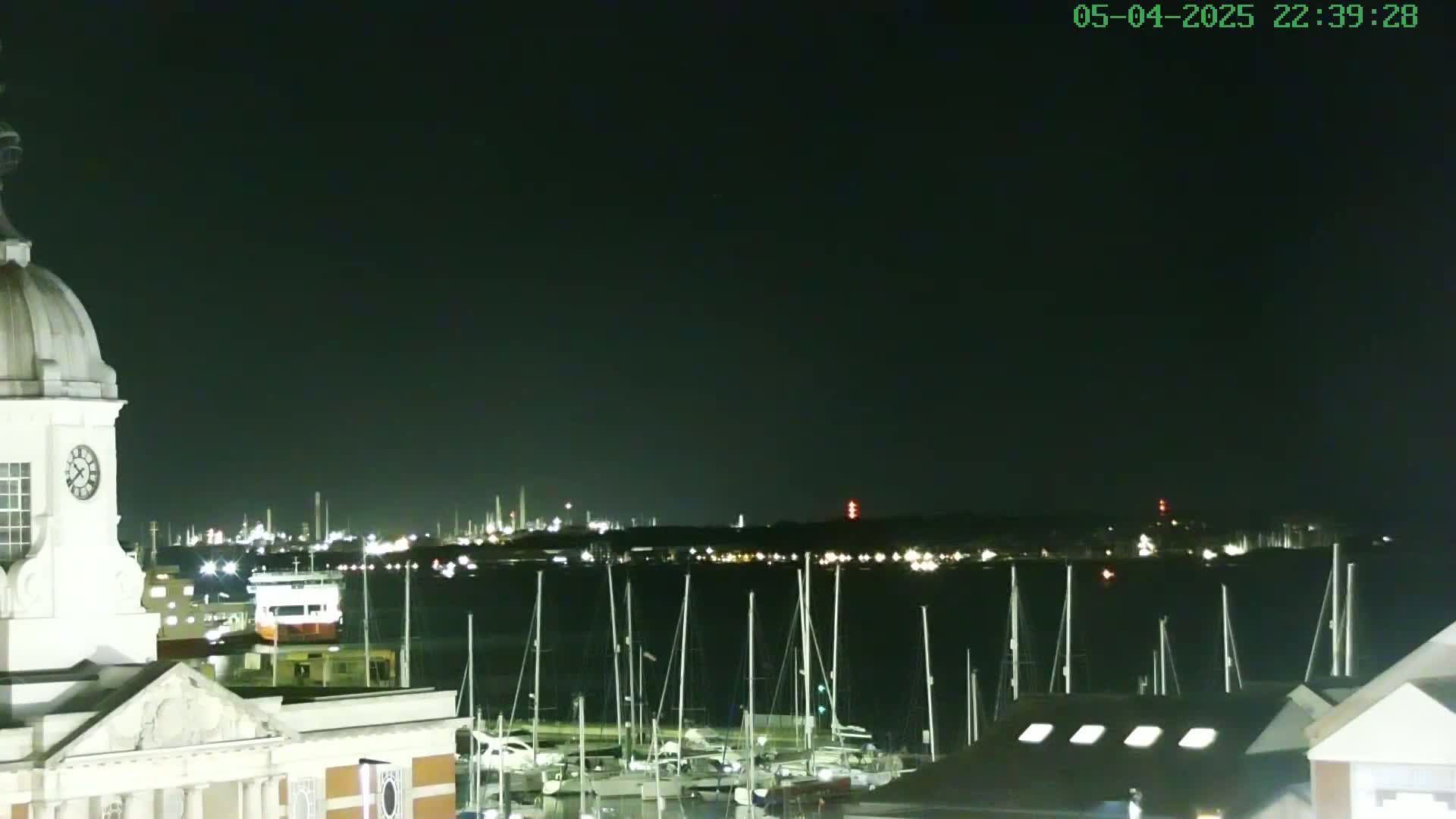 A nighttime view from a building with a clock tower shows a harbor full of sailboats and a distant industrial area lit up across the water under a clear, dark sky.