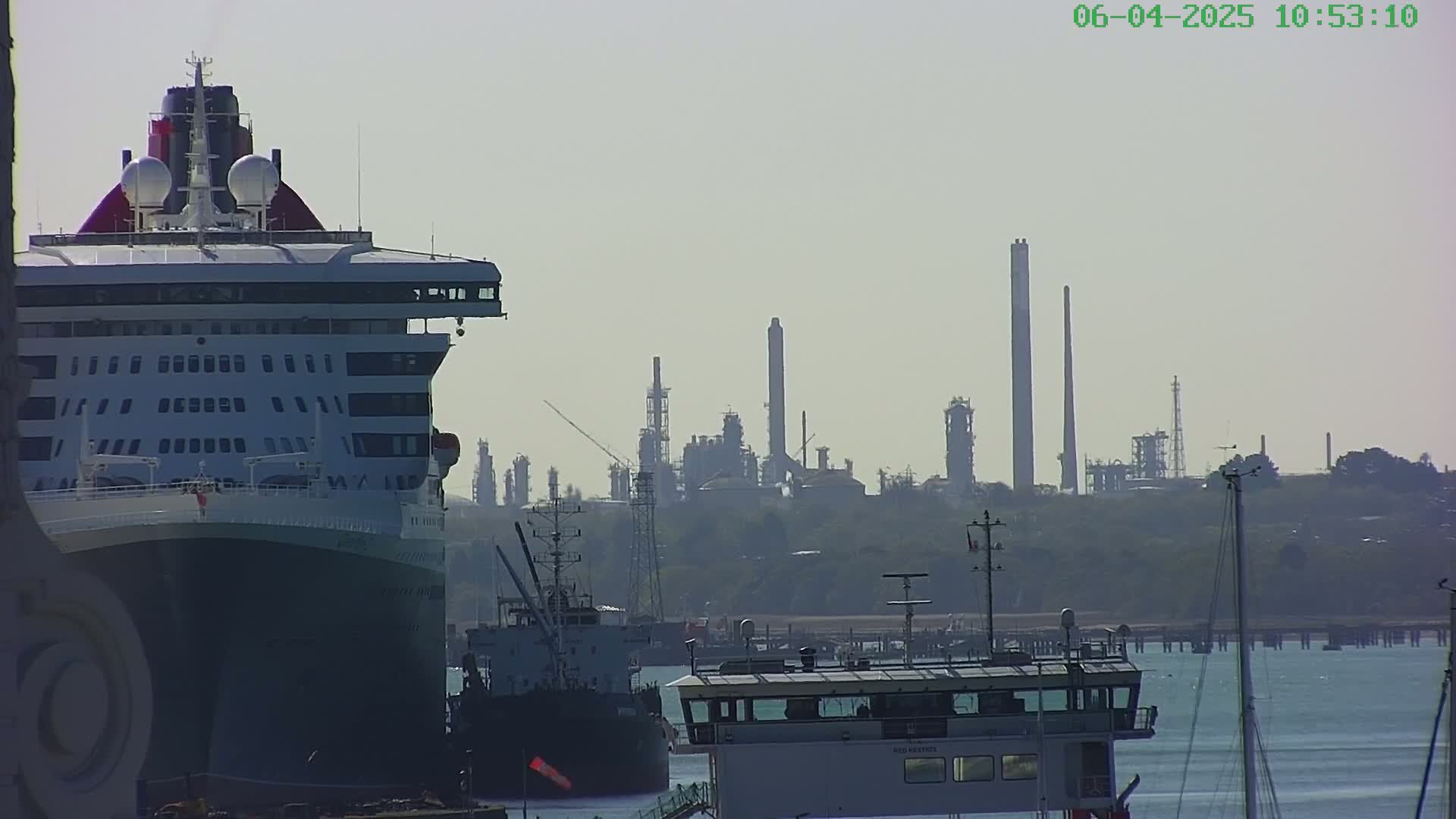 A large cruise ship is docked near an industrial refinery on a hazy day.