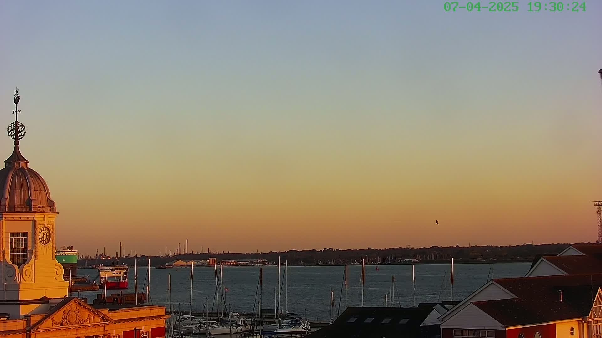 A waterfront scene at sunset shows a domed building with a weathervane, numerous boats moored in the water, and a distant industrial skyline under a clear, orange-tinged sky.
