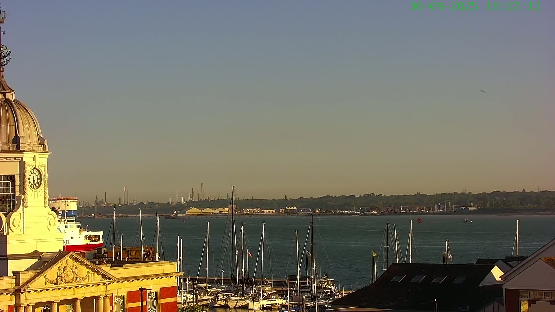 A clock tower overlooks a harbor filled with sailboats on a calm, clear day with a hazy industrial area visible in the distance.