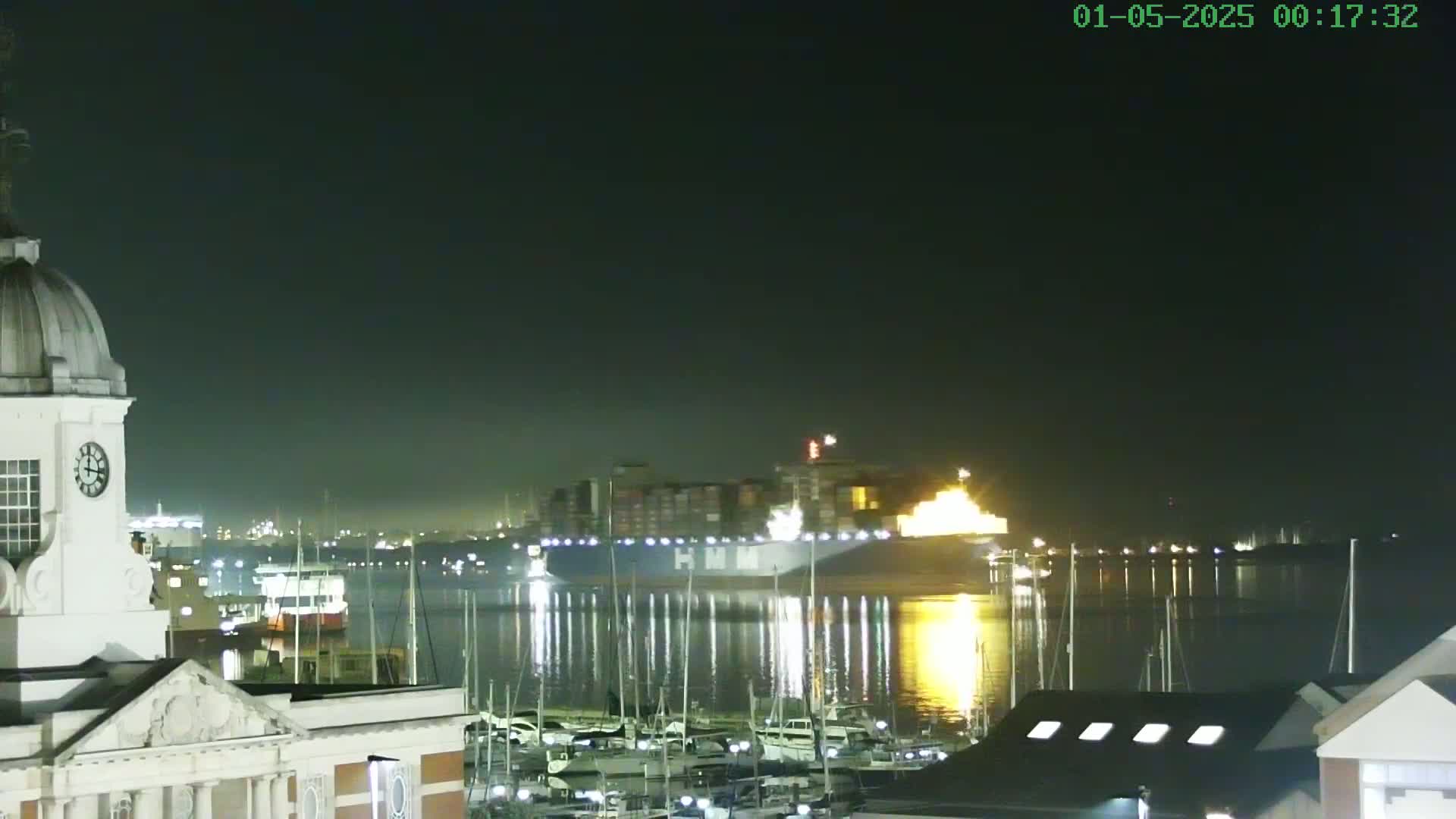 At night, a large container ship is in a harbor with many smaller boats moored nearby, under a dark, clear sky.