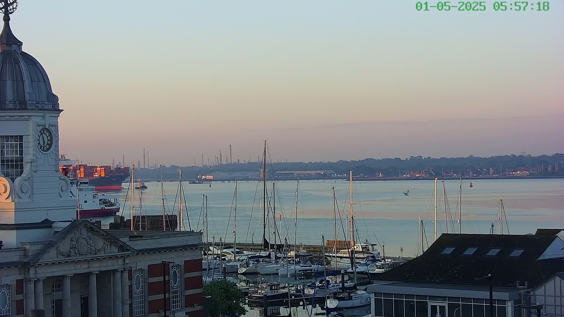 A harbor filled with sailboats at sunrise, viewed from a building with a clock tower, shows a large cargo ship and industrial structures in the distance.