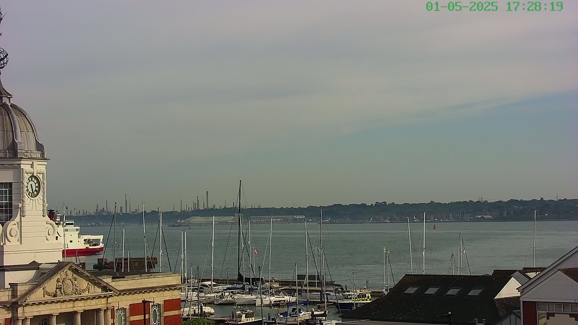 A clock tower overlooks a harbor filled with sailboats, a ferry, and a distant industrial skyline under a hazy sky.