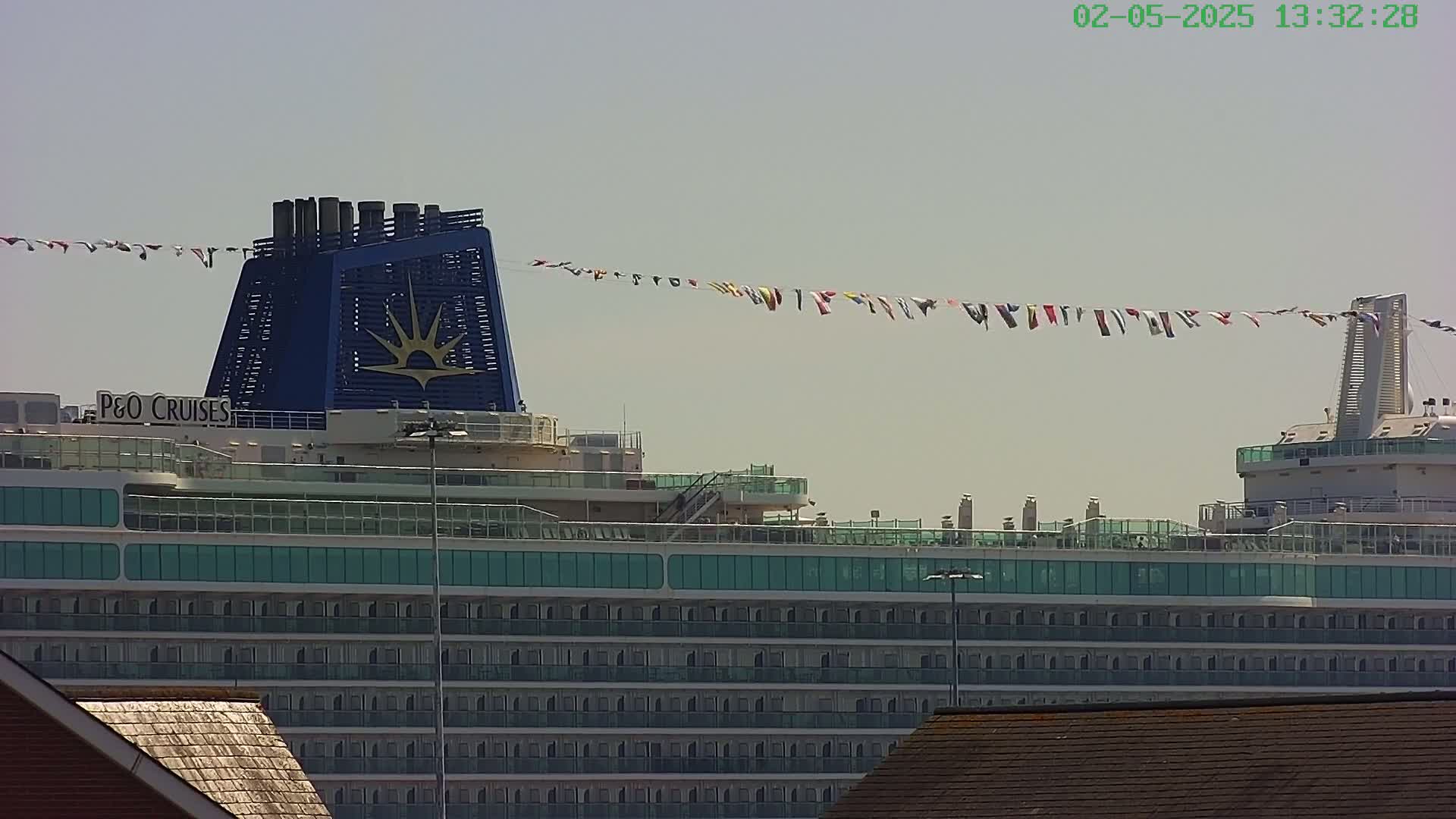 A large cruise ship with teal balconies and a blue and gold smokestack is docked under a mostly clear sky, with a string of flags strung overhead.