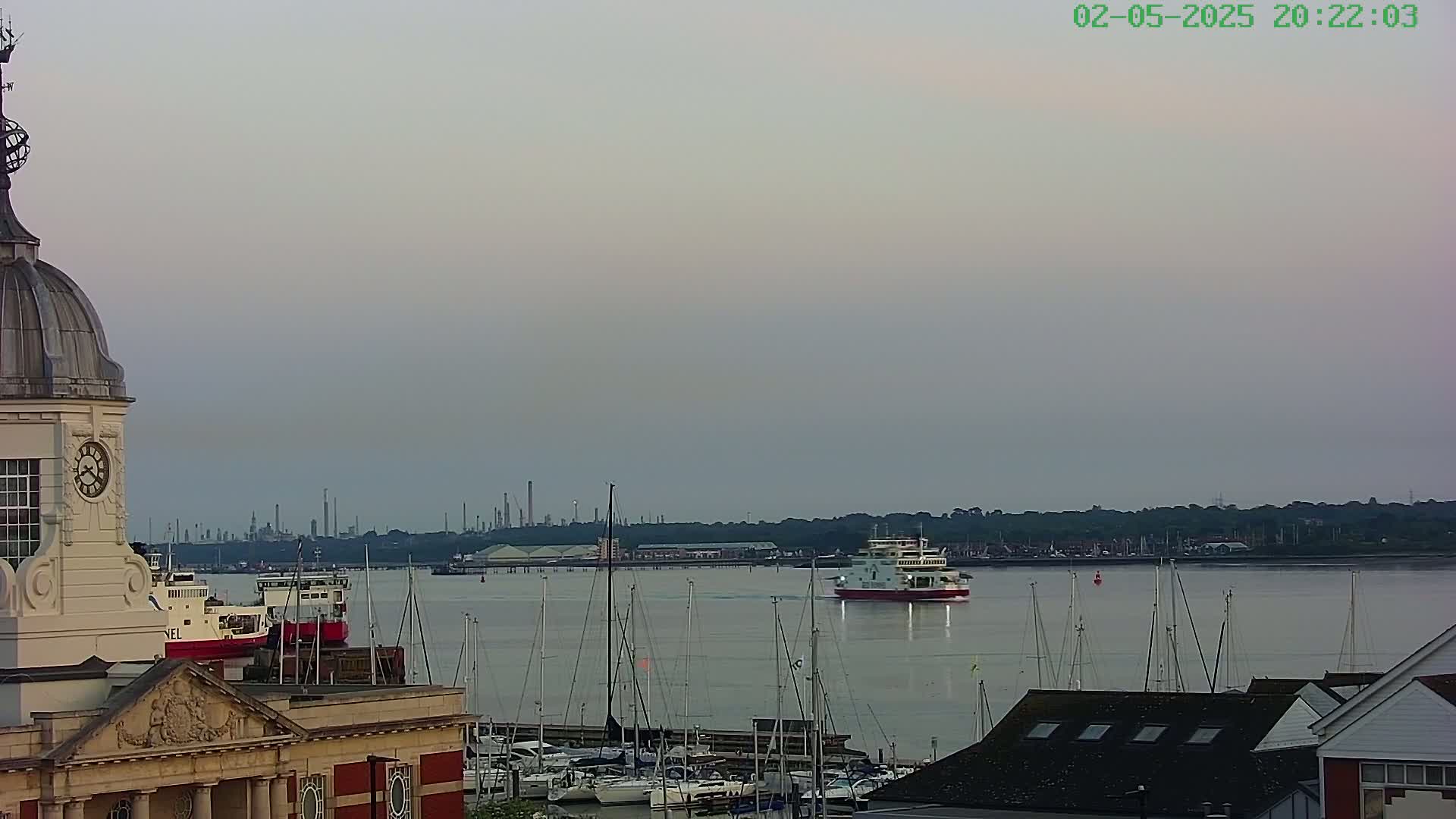 A ferry sails past a marina filled with sailboats, under a pale, overcast sky, with an industrial skyline visible in the distance.
