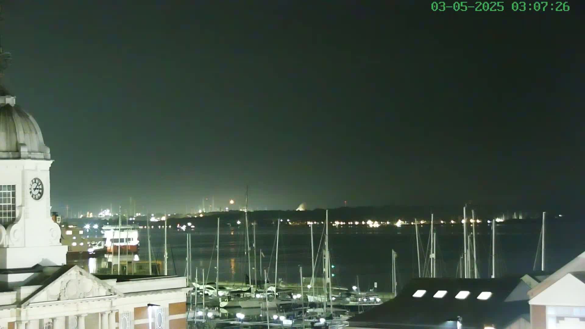 A nighttime view of a harbor filled with docked sailboats, seen from atop a building with a clock tower, under a dark sky.
