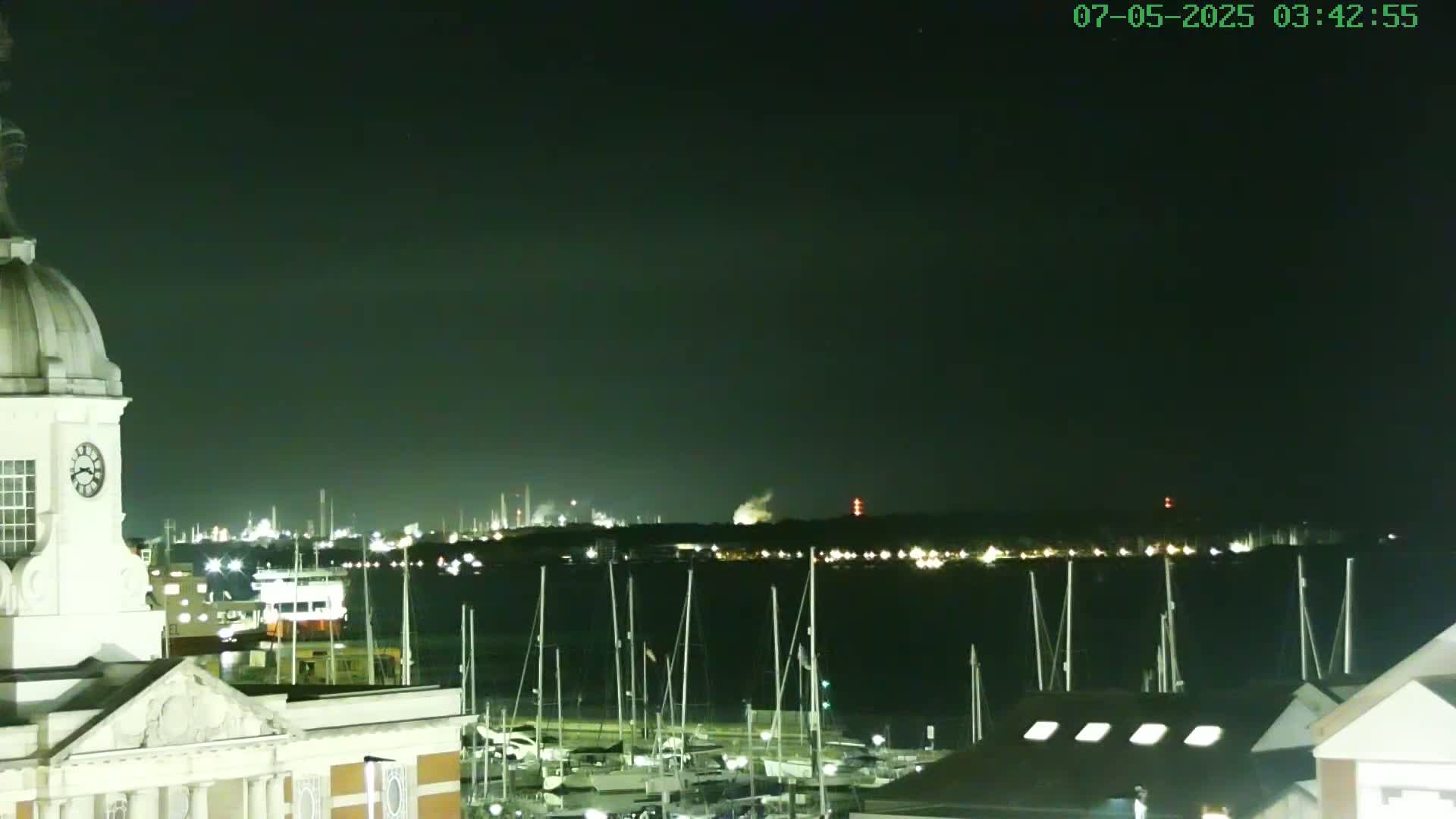 A nighttime view from a waterfront building shows a harbor filled with docked sailboats, with an industrial area illuminated in the distance under a dark sky.