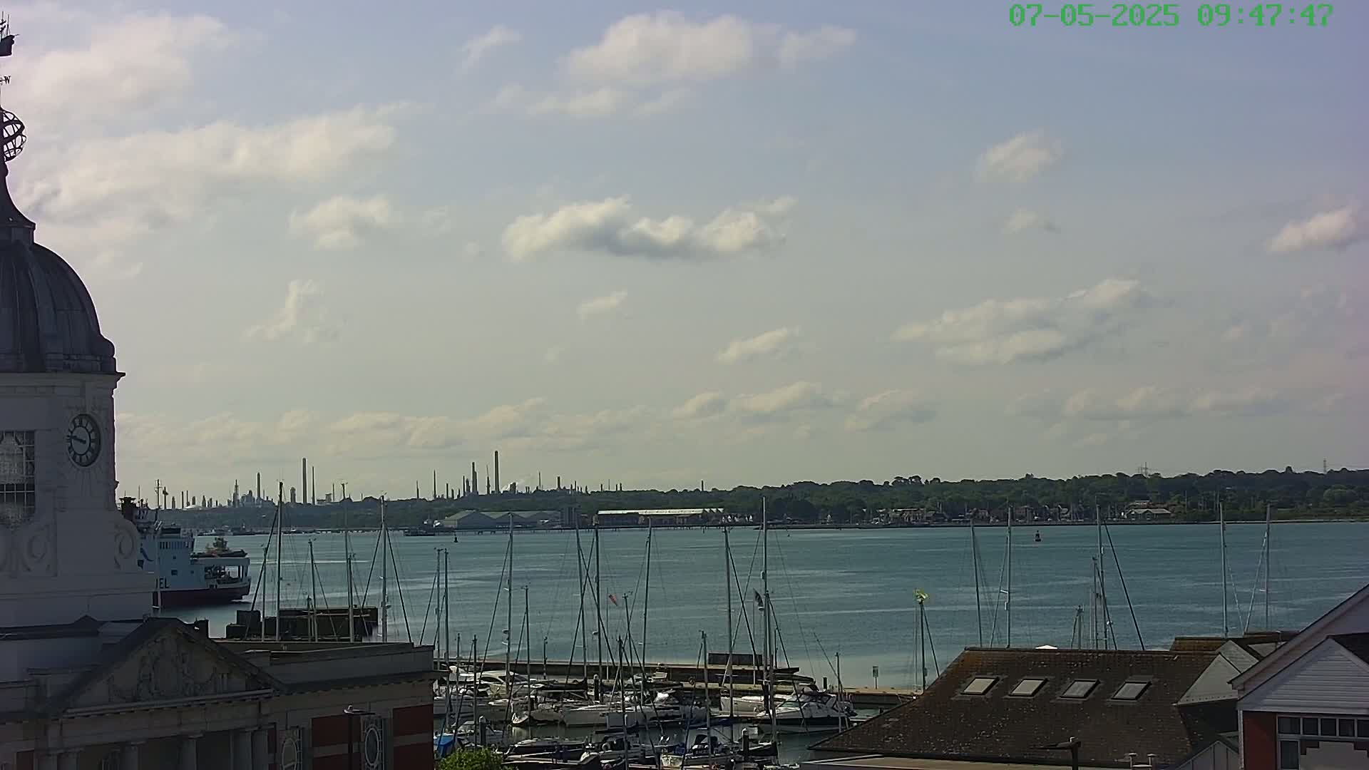 A partly cloudy sky overlooks a marina filled with sailboats, a ferry, and an industrial plant in the distance.
