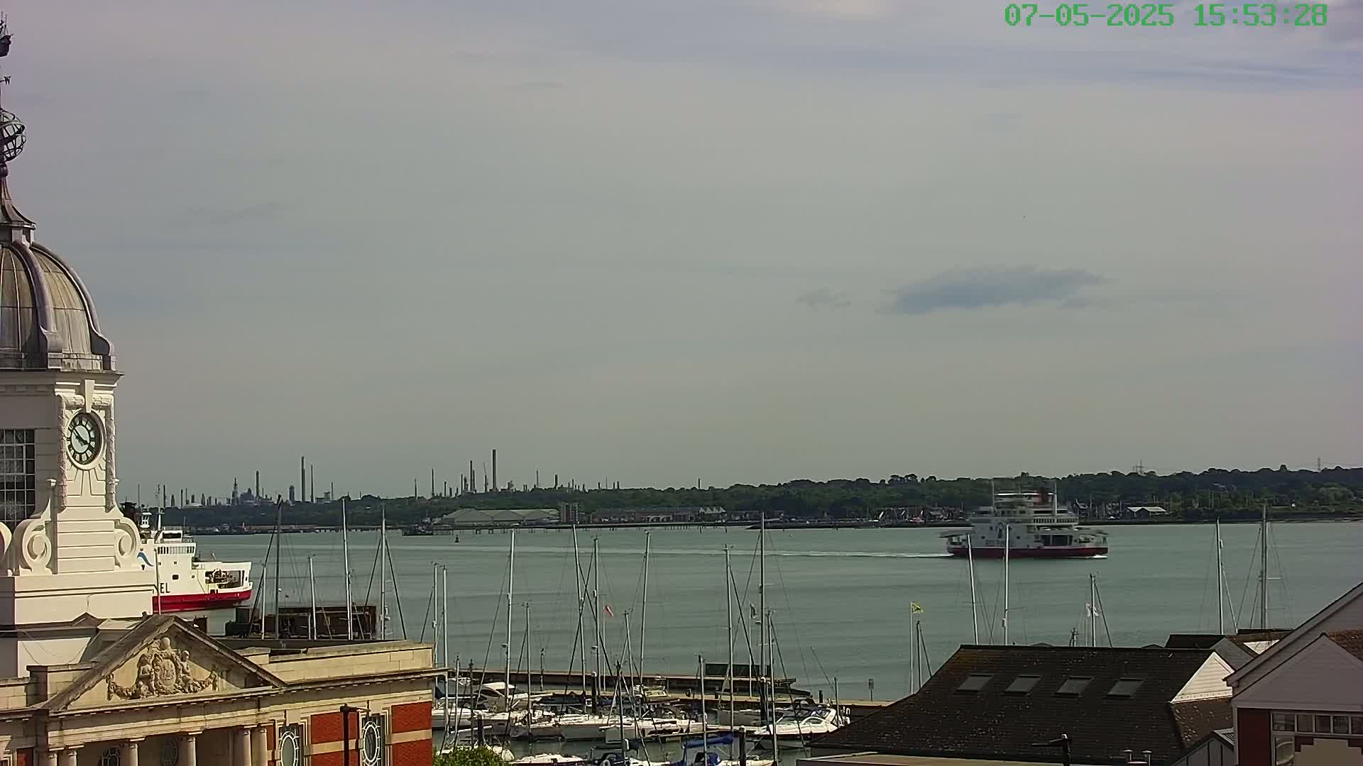 A clock tower overlooks a harbor filled with sailboats, a ferry, and a distant industrial skyline under a partly cloudy sky.