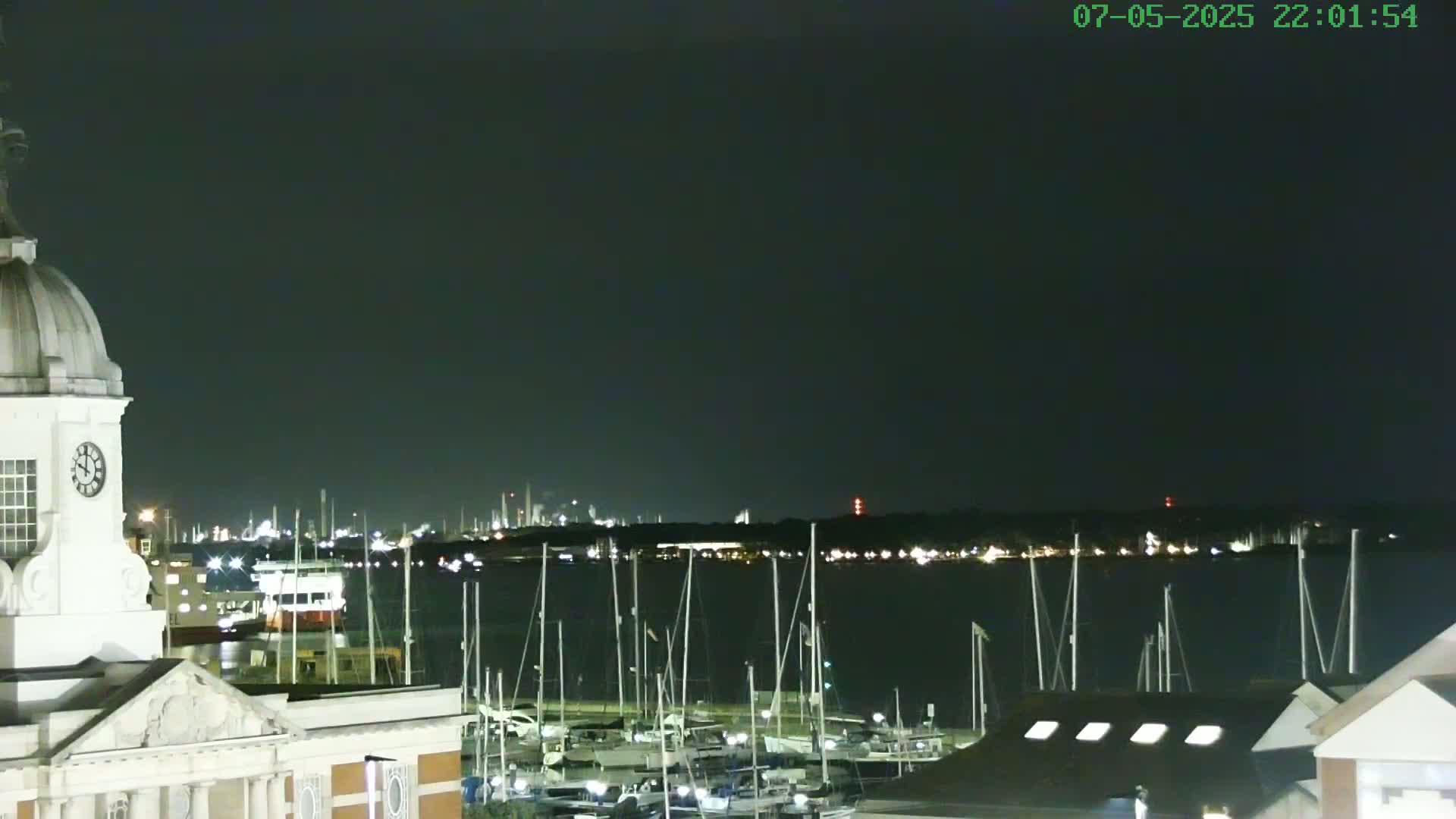 A nighttime view of a harbor filled with docked sailboats, seen from a vantage point near a clock tower and building, under a dark, clear sky.