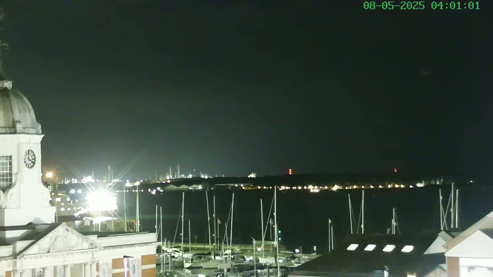 A nighttime view from atop a building with a dome and clock shows a dark body of water with many masts of sailboats docked, a distant industrial area with lights, and a clear, dark sky.