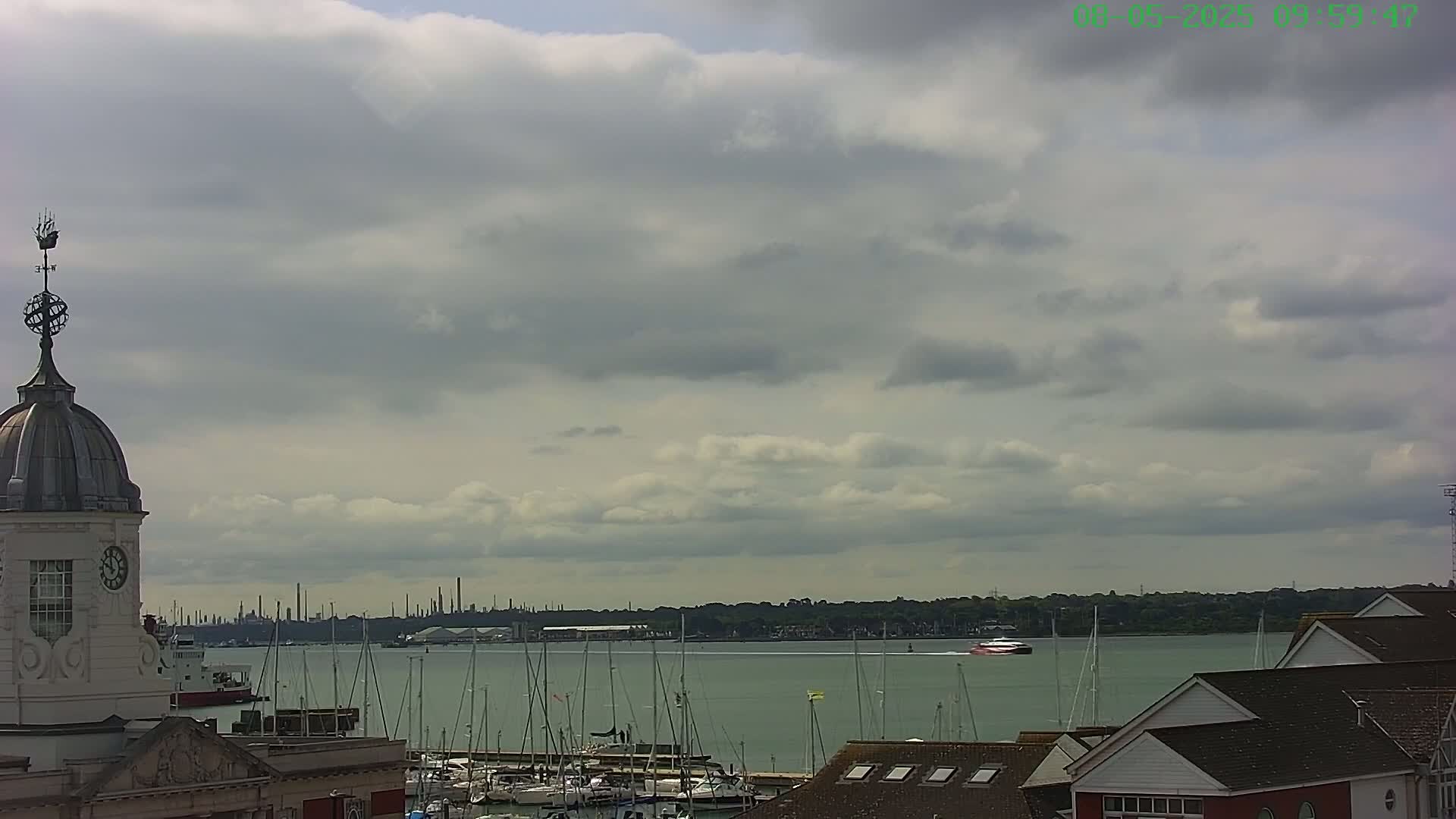 A partly cloudy sky overlooks a harbor with numerous sailboats and a distant industrial skyline.