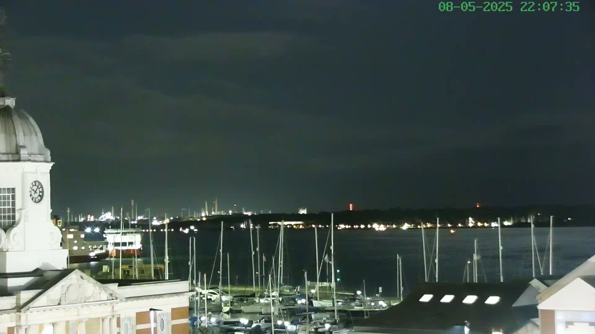 A nighttime view from a building with a clock tower overlooking a harbor filled with docked sailboats and a distant, illuminated shoreline under a dark, cloudy sky.