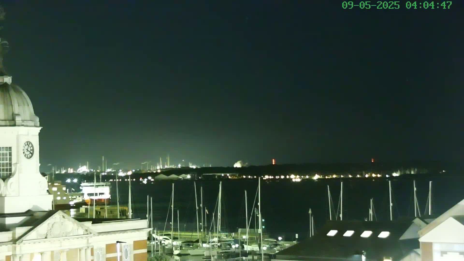 A nighttime view from atop a building shows a harbor filled with boats and a distant industrial area illuminated under a clear, dark sky.