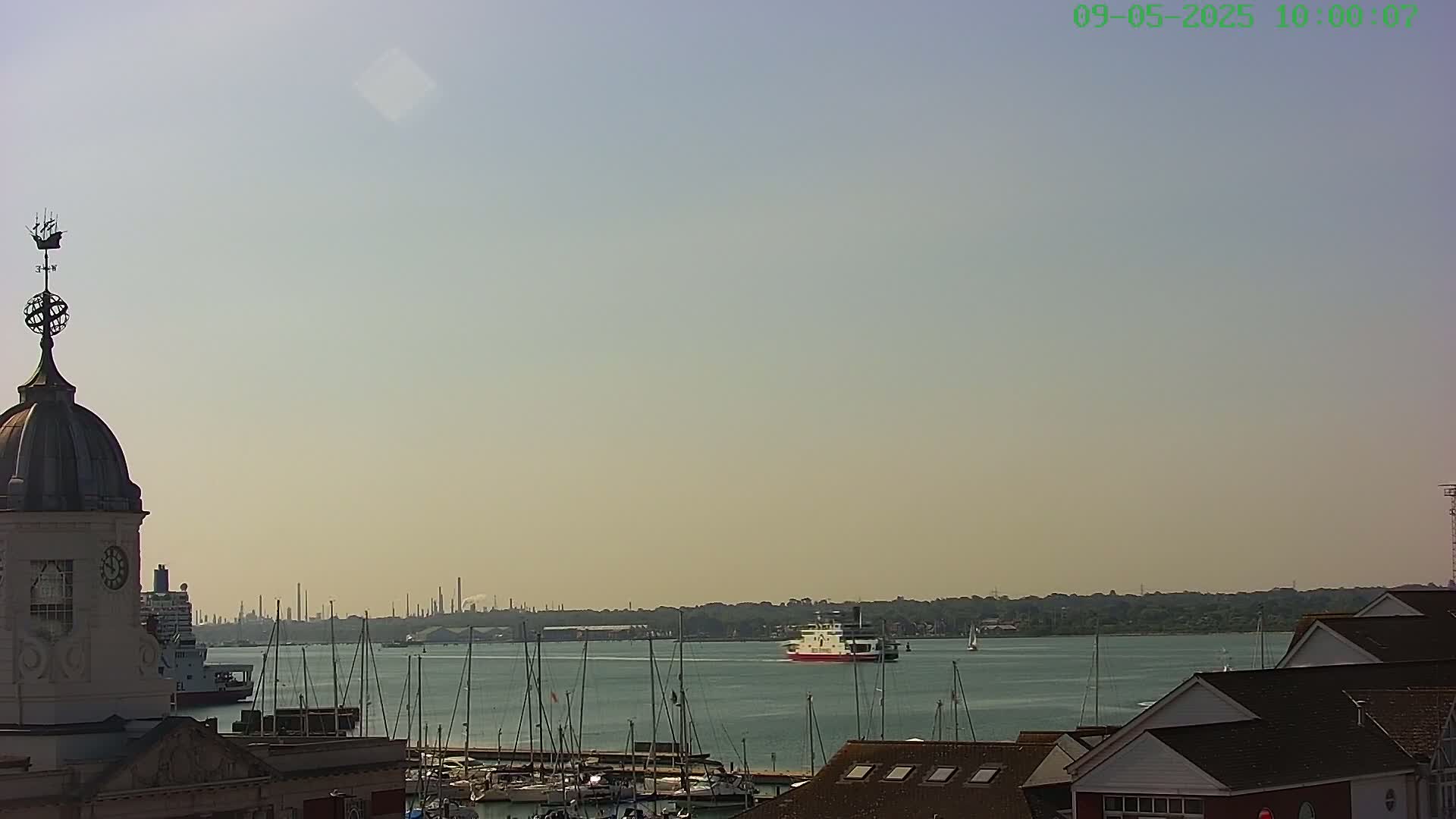 A ferry passes by a harbor filled with sailboats under a hazy, sunny sky, with a clock tower visible in the foreground and an industrial skyline in the distance.