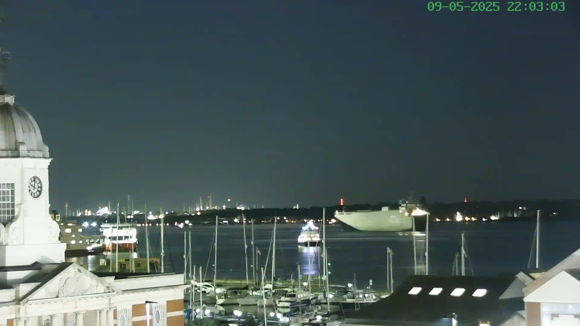A nighttime view of a harbor with numerous boats and a large ship in the distance under a dark, clear sky.