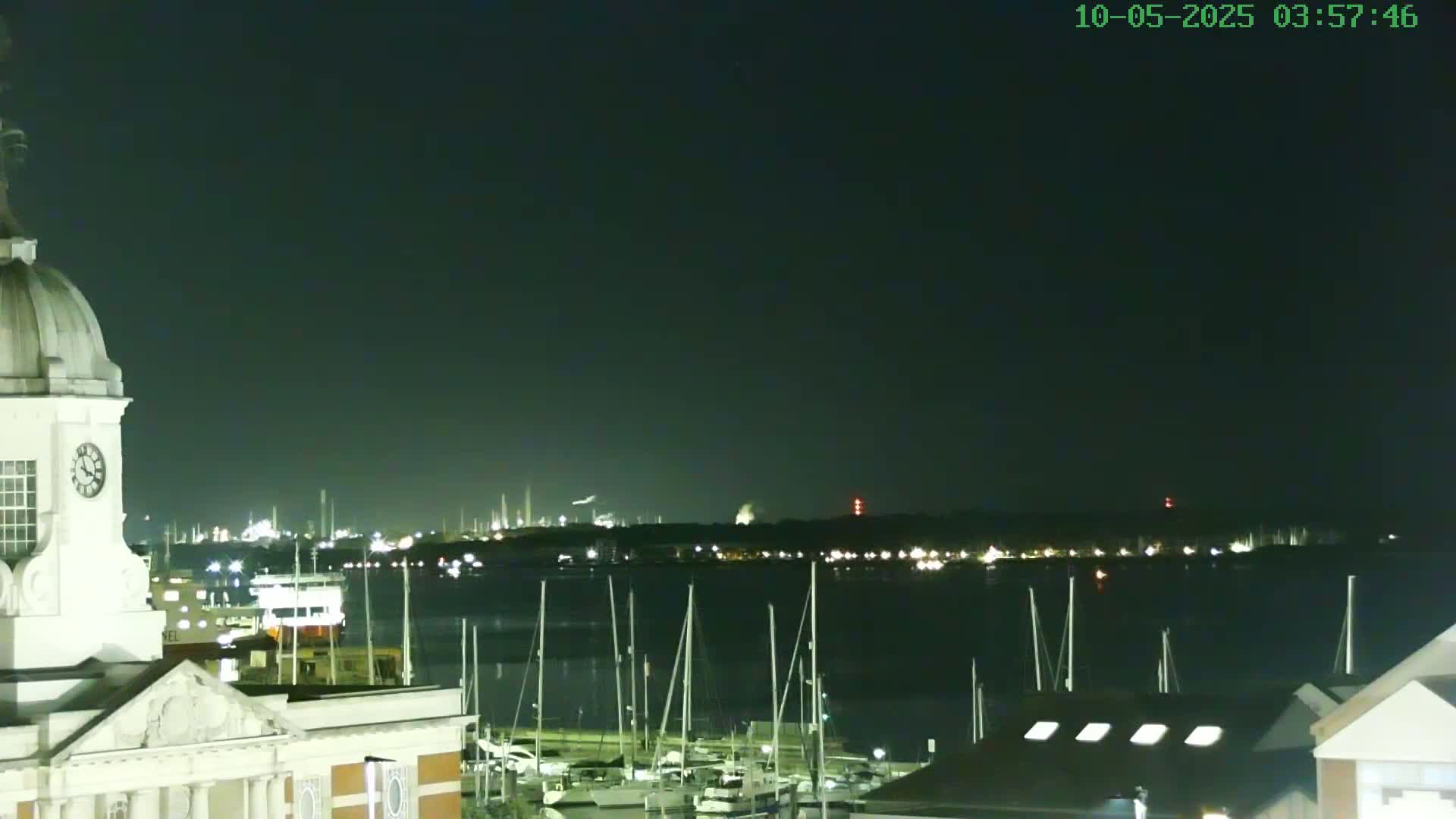 A nighttime view from atop a building shows a calm body of water with numerous docked sailboats, a distant illuminated cityscape, and a clock tower in the foreground under a clear, dark sky.