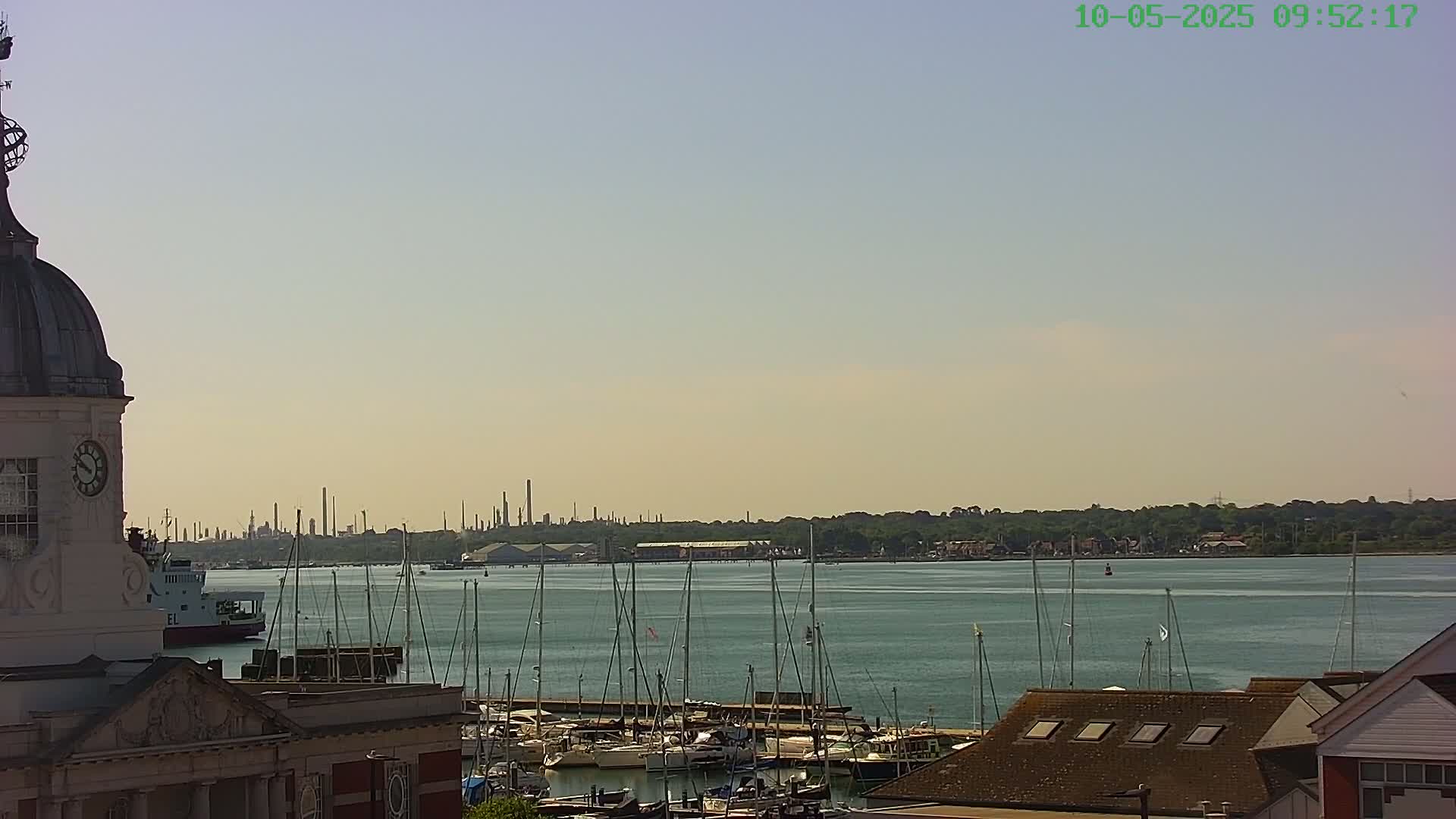 A view from a clock tower shows a calm body of water with numerous sailboats docked, a distant industrial skyline, and hazy skies.