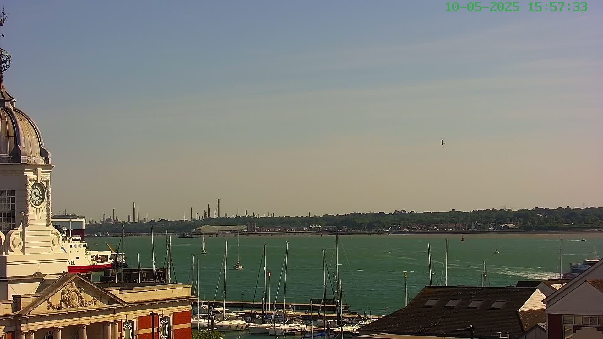 A clock tower overlooks a harbor filled with sailboats on a sunny day, with a refinery visible in the distance.
