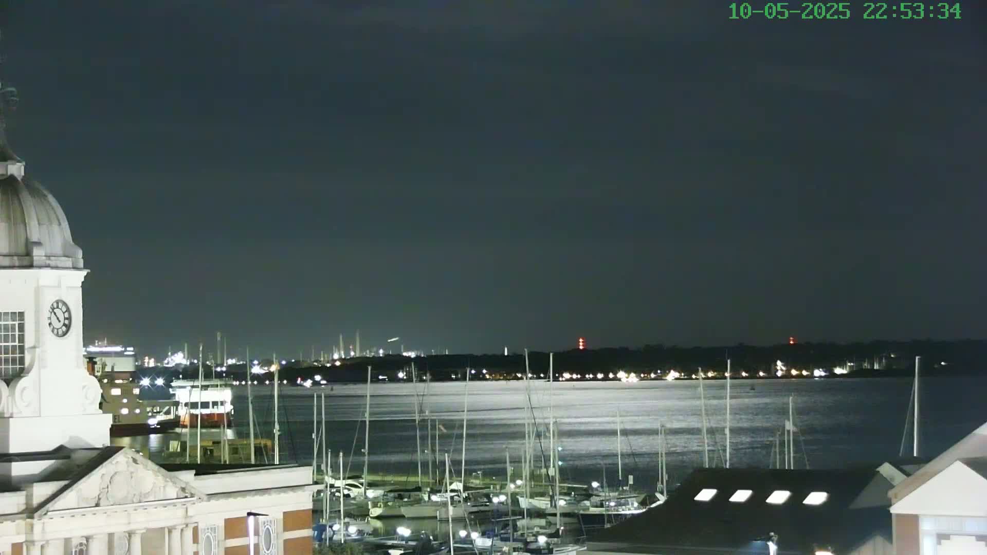 A nighttime view from atop a building shows a calm body of water with many sailboats moored in a marina, a city skyline in the distance, and a clock tower in the foreground under a dark sky.
