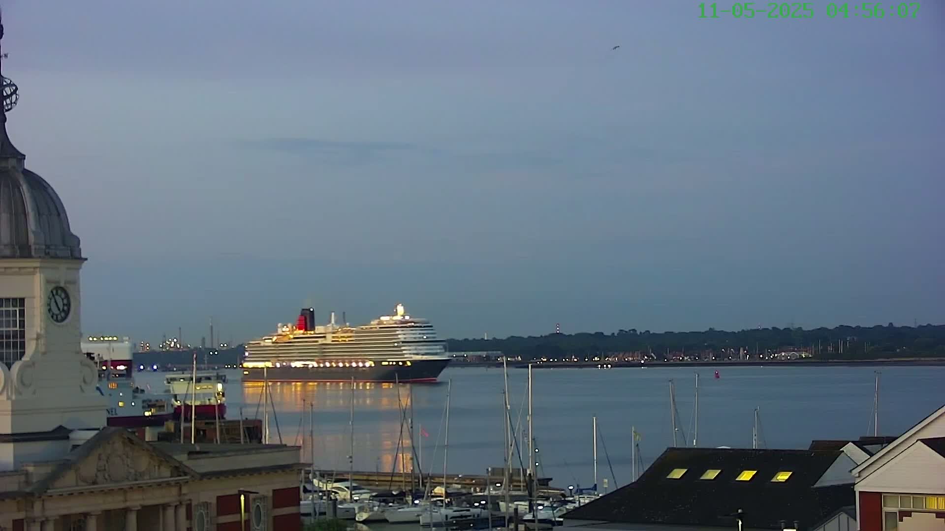 A large cruise ship is in the harbor at twilight, with many smaller boats moored nearby and a building with a clock tower in the foreground.