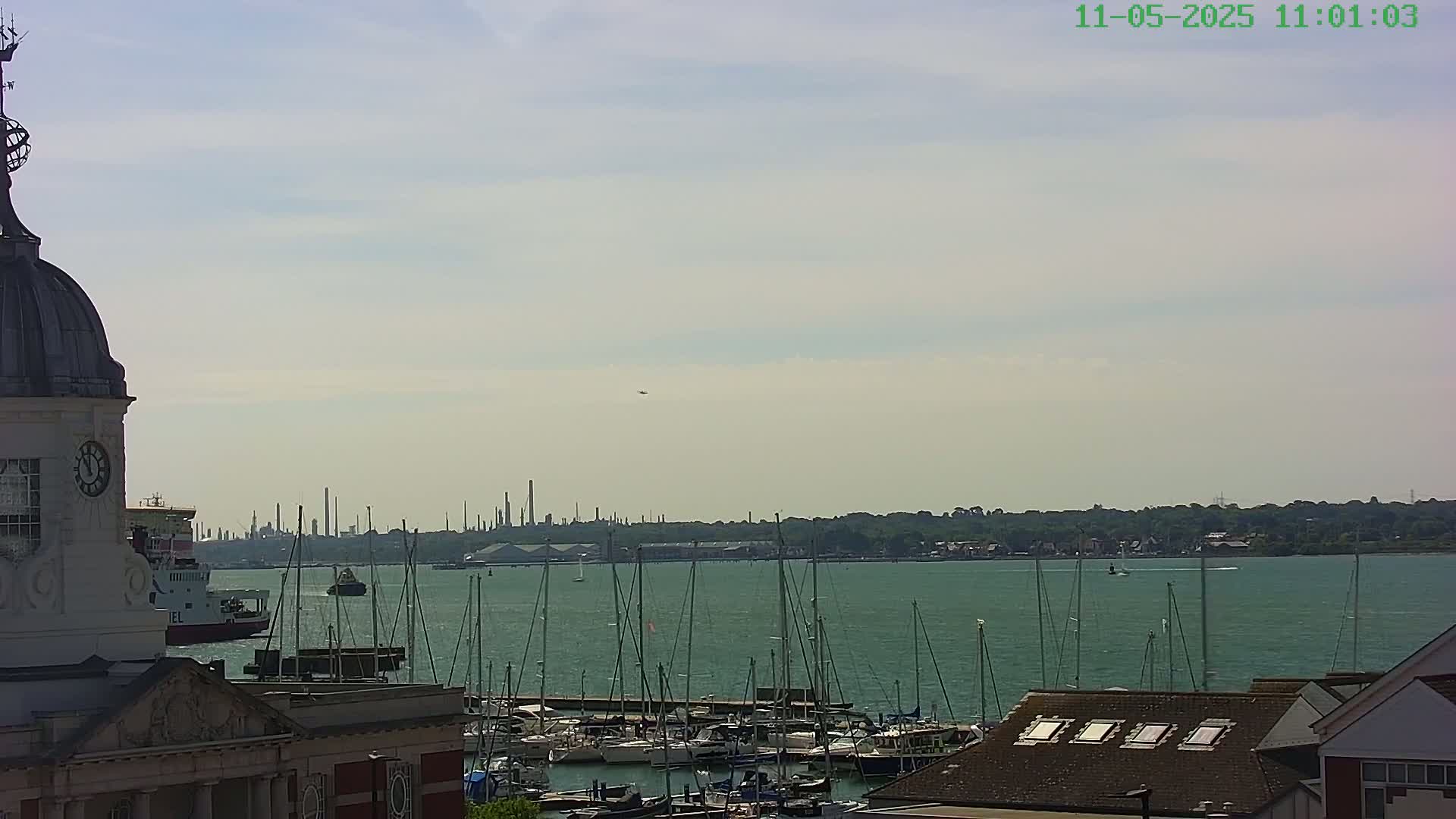 A partly cloudy day shows a harbor filled with sailboats, a distant industrial area, and a portion of a clock tower building.