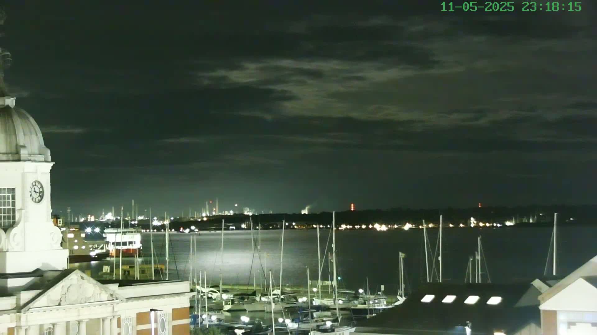 A nighttime view of a harbor filled with docked sailboats, overlooked by a clock tower on a building, under a partly cloudy sky.