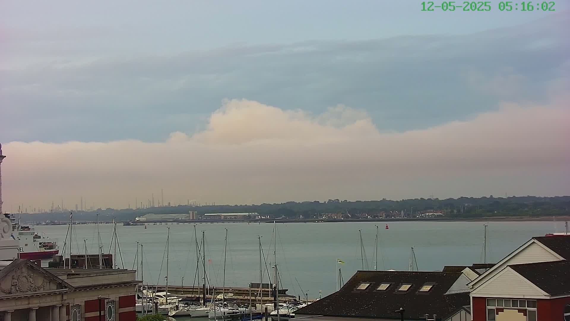A calm body of water with numerous sailboats docked, a distant industrial shoreline, and a partly cloudy sky.