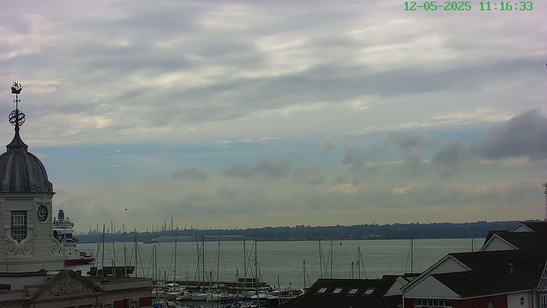 A partly cloudy sky overlooks a harbor filled with sailboats and a distant industrial skyline.